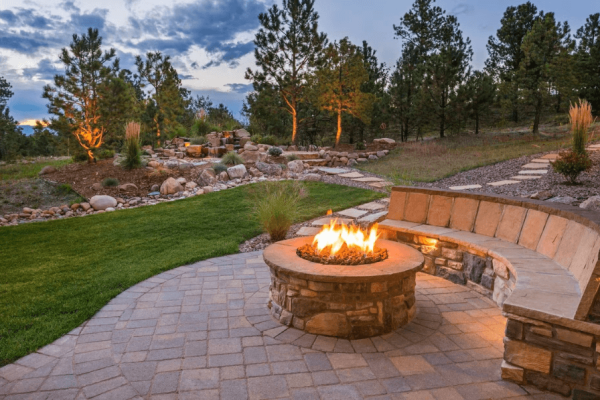 A photo of a large yard with a stone patio, bench, and firepit. They are surrounded by green grass and landscaping features with natural stone and shrubs.