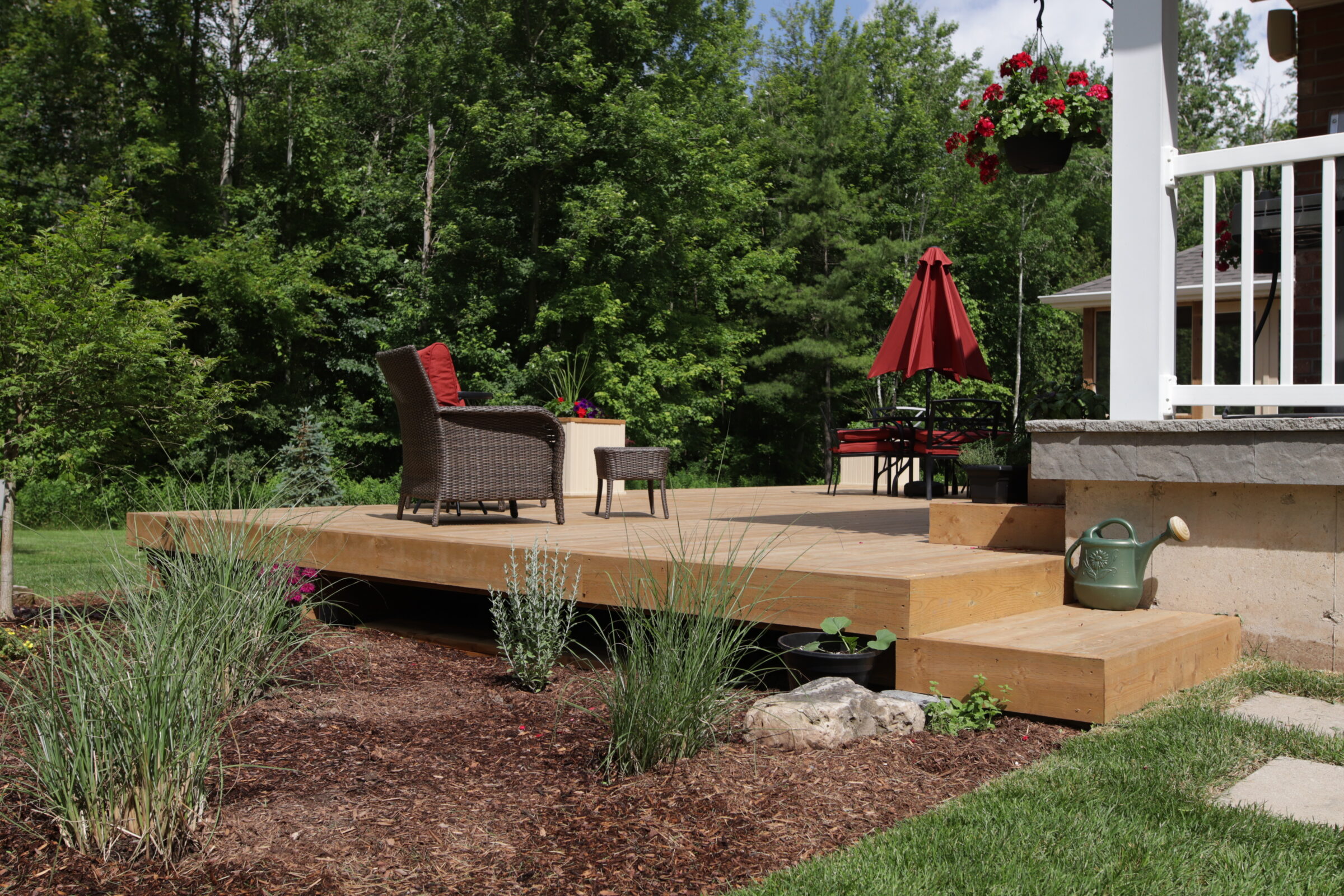 A wooden deck with wicker furniture and a red umbrella overlooks a lush garden and forest. Steps lead up to a house with a white railing.