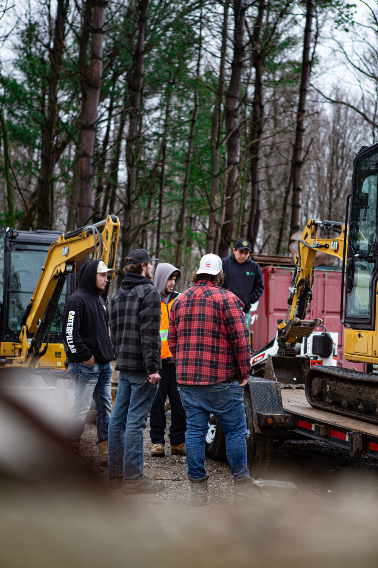 A group of people, some in high-visibility vests, gather near a yellow excavator in a wooded area, possibly discussing work-related matters.
