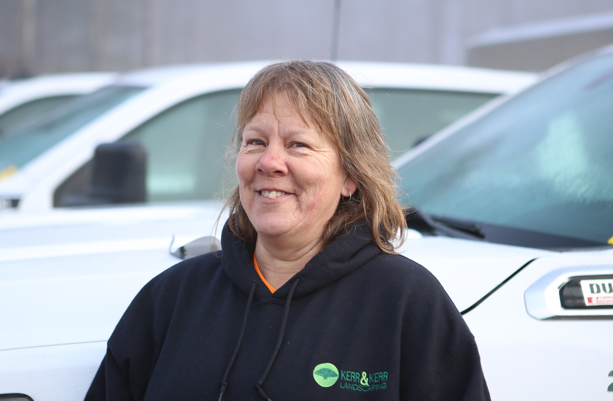 A smiling person in a black hoodie with a logo stands outdoors with white vehicles in the blurred background on an overcast day.