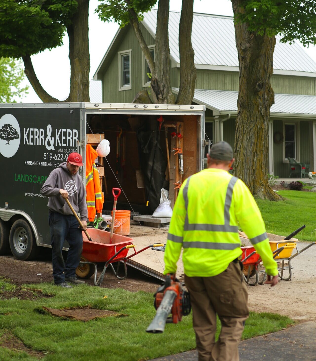 Two people in high-visibility vests are working near a landscaping trailer parked by a house amid greenery. Lawn care tools and equipment are visible.