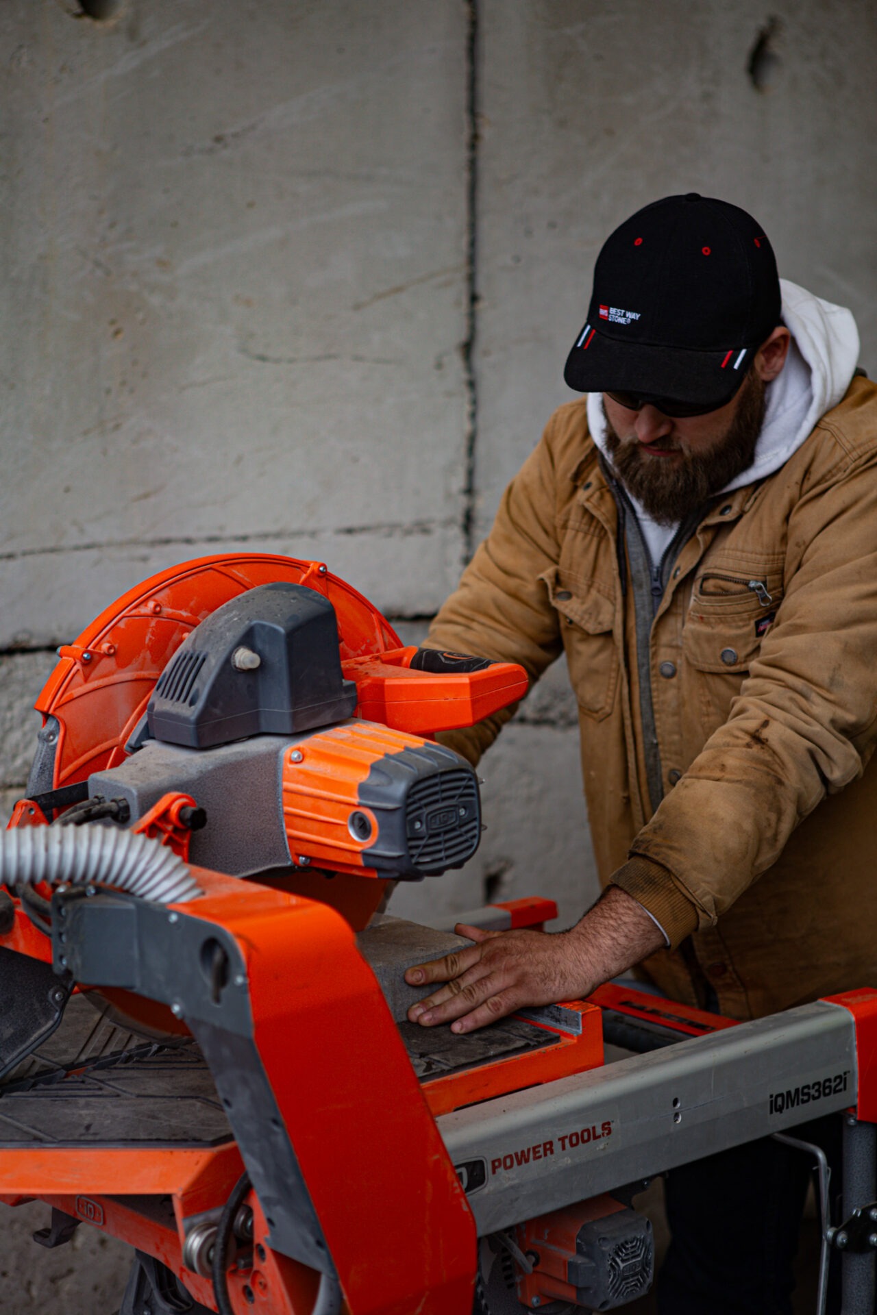 A person wearing a cap and a jacket operates an orange and gray concrete saw, with a focus on precision and safety.