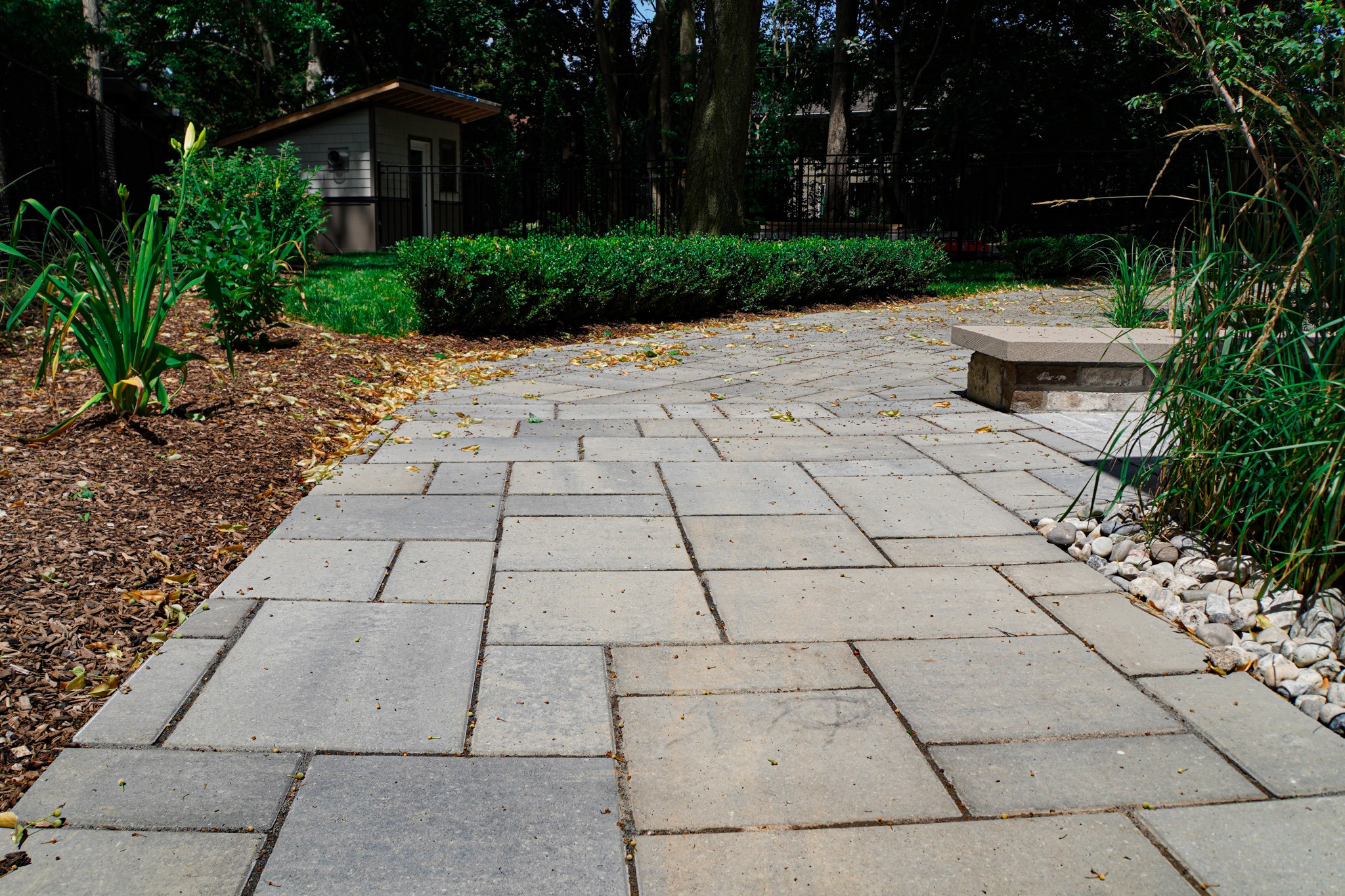 Stone pathway leading through a landscaped garden with plants and mulch, towards a small building with a black fence behind it under a cloudy sky.