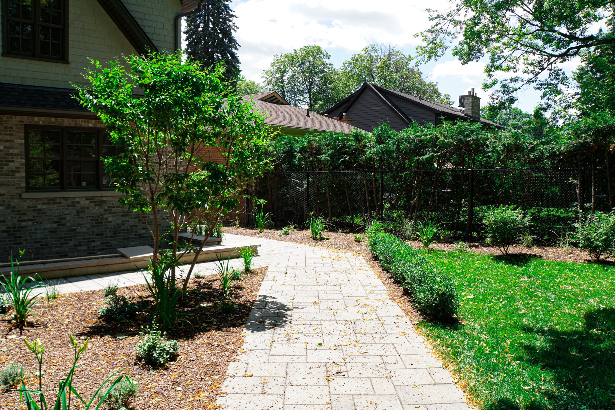 A backyard using modern landscaping ideas such as a stone pathway, lush greenery, a bench near the house, and a chain-link fence under a sunny sky.