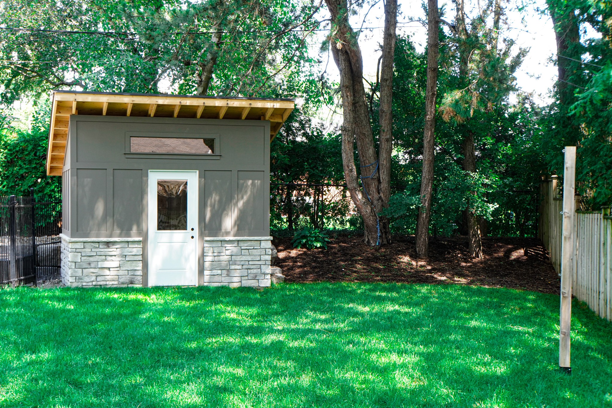 A small modern shed with gray siding and stone foundation sits on a lush green lawn surrounded by trees behind a black metal fence.