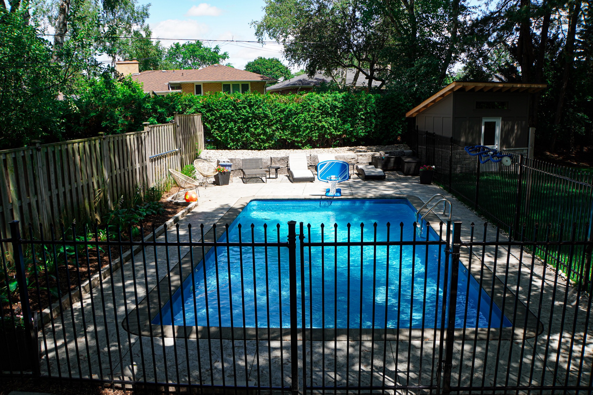 A backyard with a rectangular in-ground pool surrounded by a metal fence, patio with chairs, trees, and a small shed under a sunny sky.