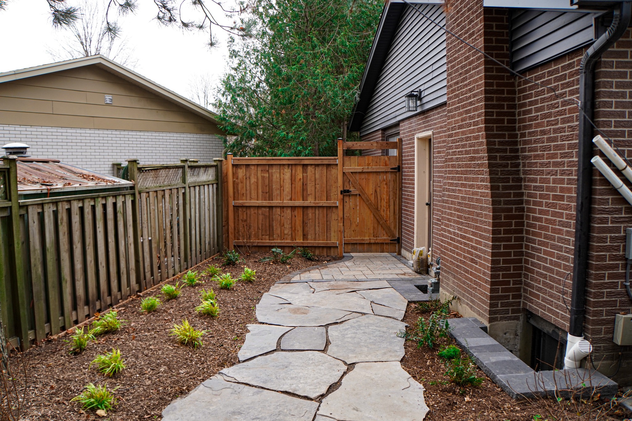 A stone pathway leads to a wooden gate between houses with brick exteriors, under a cloudy sky, alongside landscaped beds with young plants.