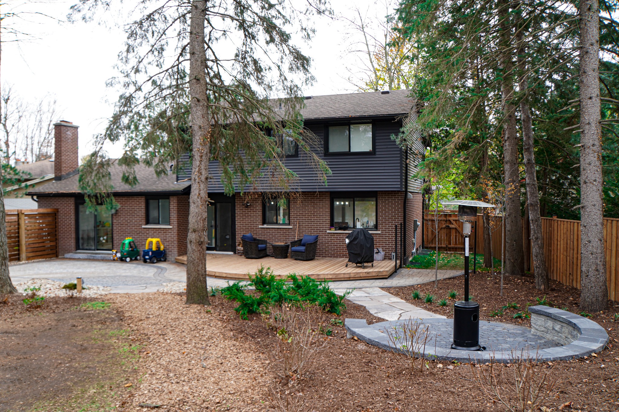 This image shows a two-story house with dark siding and brick exterior, surrounded by trees, with a wooden deck and a children's toy car outside.