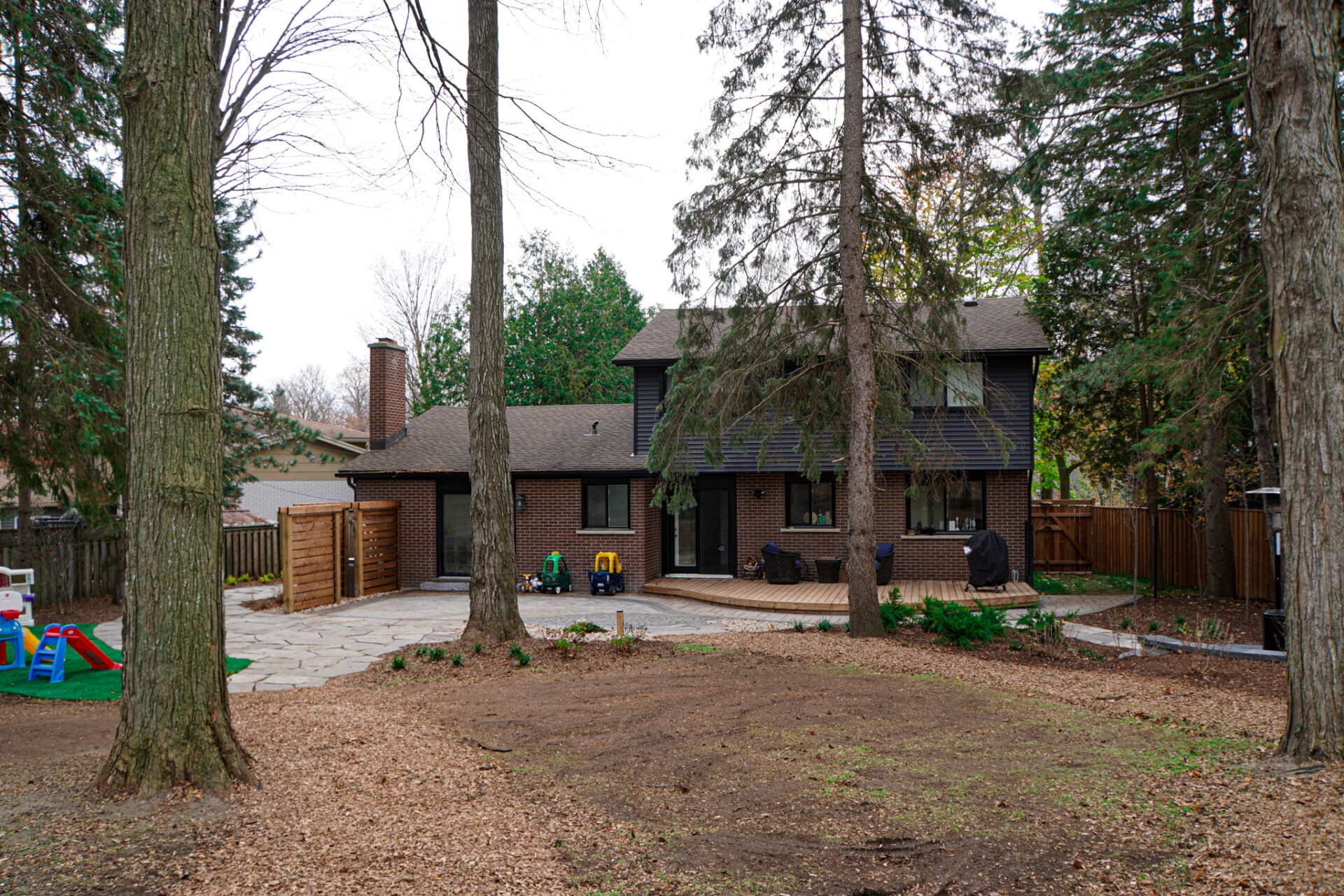 A two-story brick house with a dark roof, surrounded by trees, featuring a patio area, a wooden fence, and children's play equipment in the yard.