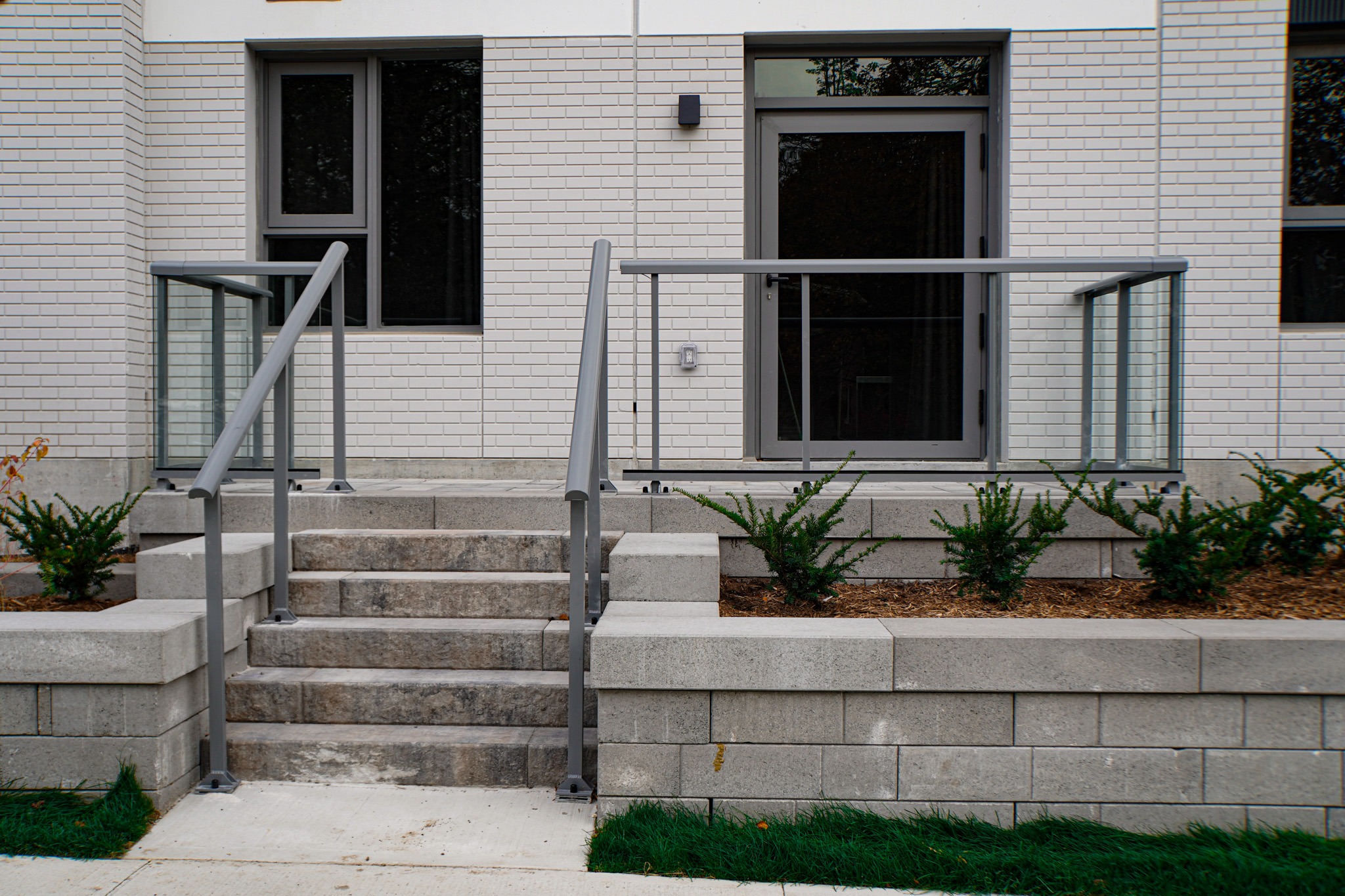 A set of concrete steps leads to a door flanked by windows on a white brick building, with metal handrails and small shrubs planted nearby.