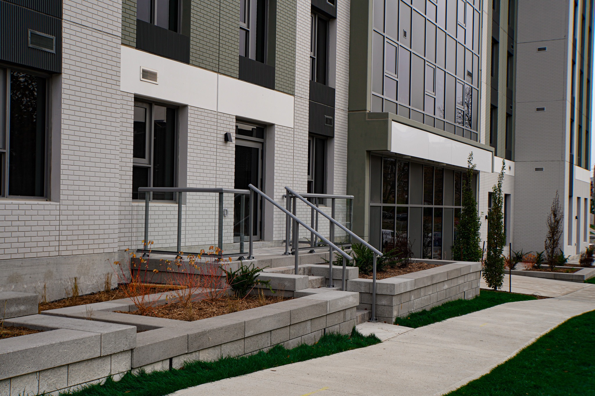 The image shows a modern building with white brick walls, large windows, and a landscaped front with steps and metal handrails.