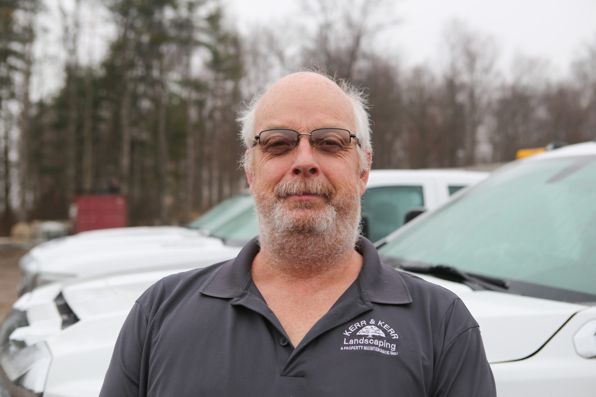 A person with glasses and a beard stands in front of white vehicles, wearing a black polo shirt with a landscaping company logo.