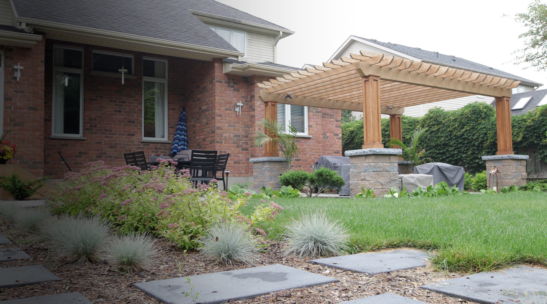 This image shows a well-landscaped front yard with a brick house, pergola, green lawn, stone pathway, and various plants and shrubs.