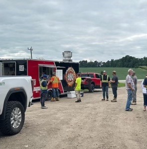 A group of people queues at a red food truck in a rural setting with vehicles and open fields in the background under an overcast sky.