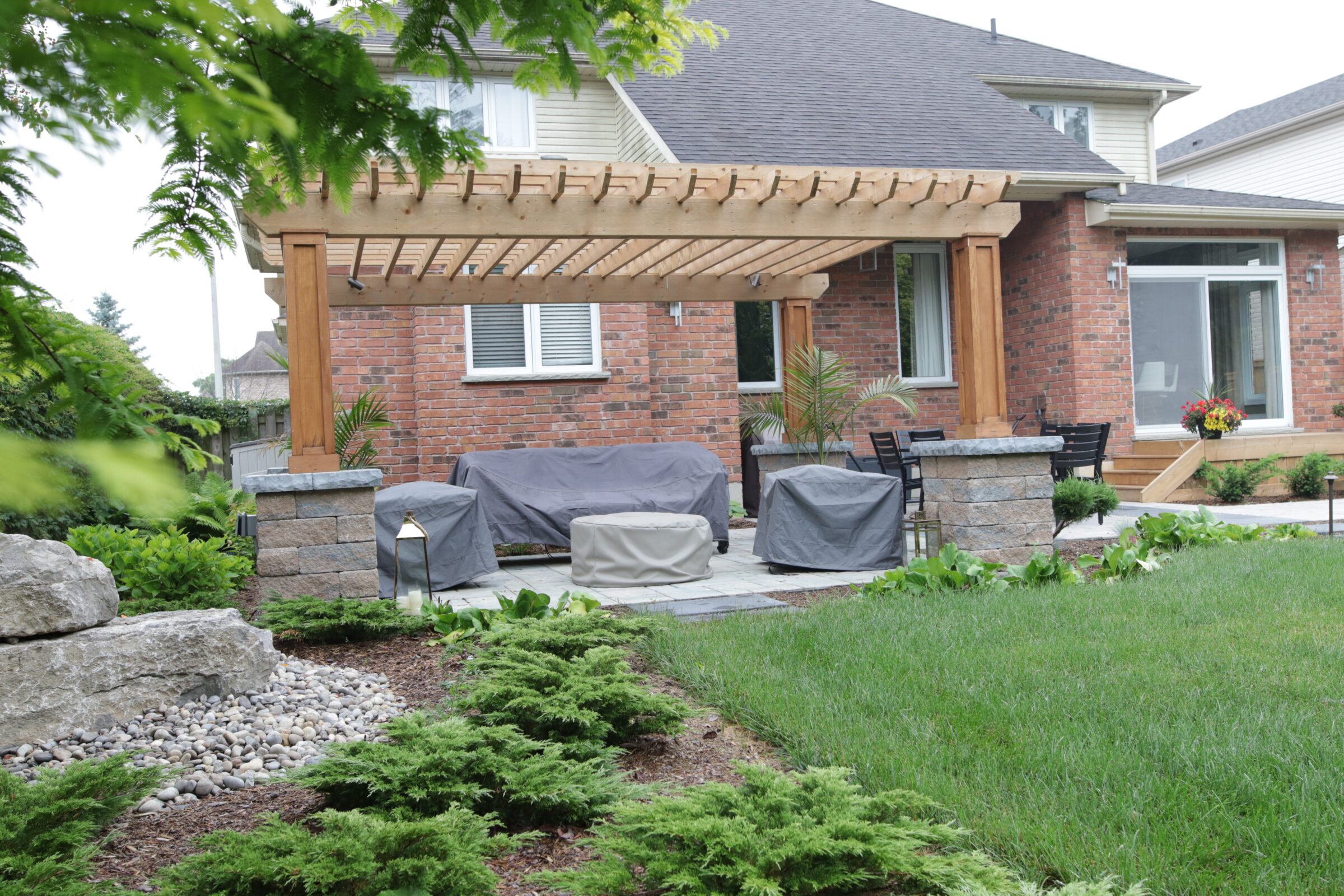 This image shows a residential backyard with a covered patio, landscaped garden, outdoor furniture covered with protective cloths, and a well-maintained lawn.