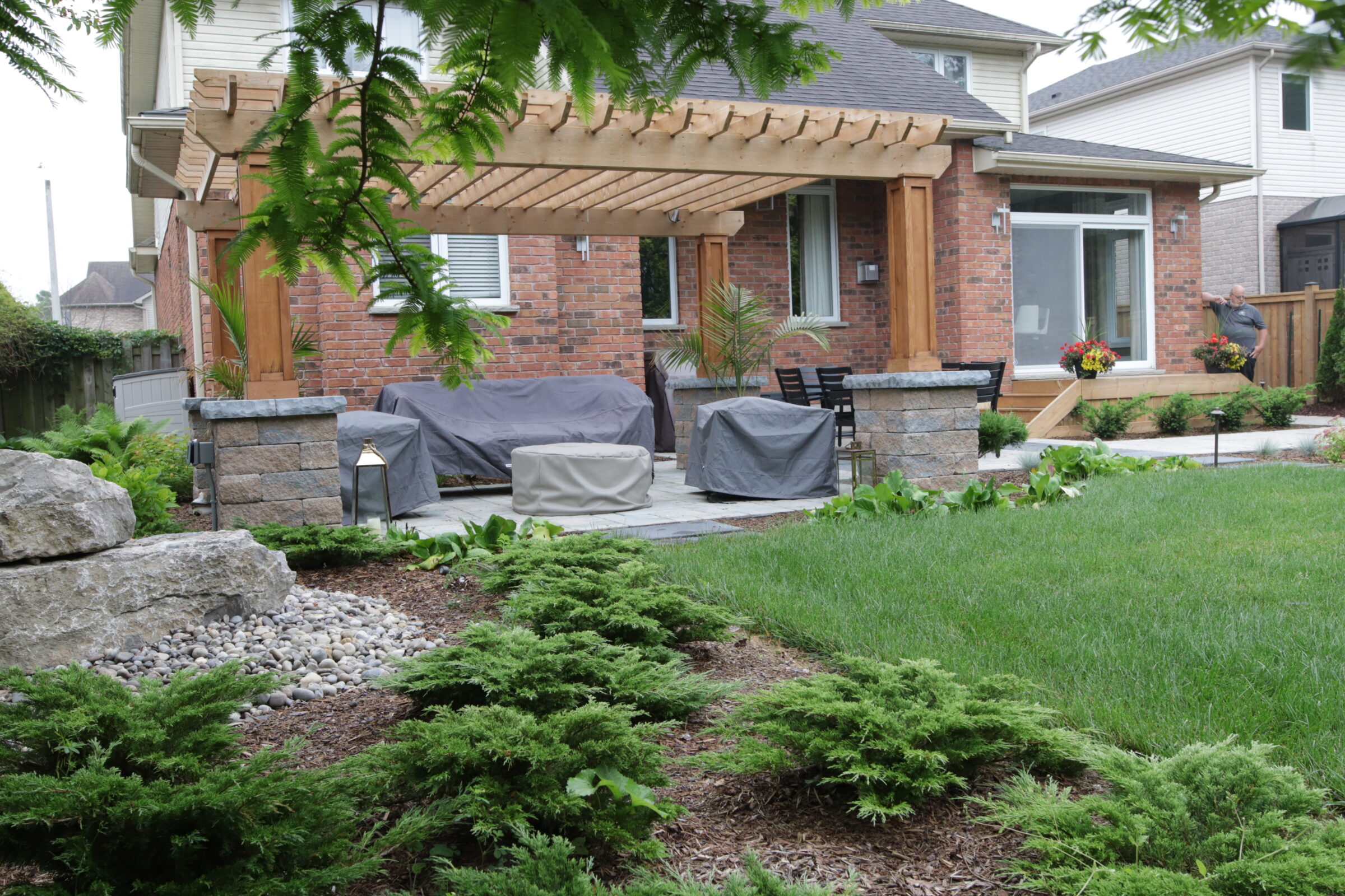 A well-kept garden with a pergola, outdoor furniture with covers, and a brick house. A person is in the background tending to plants.