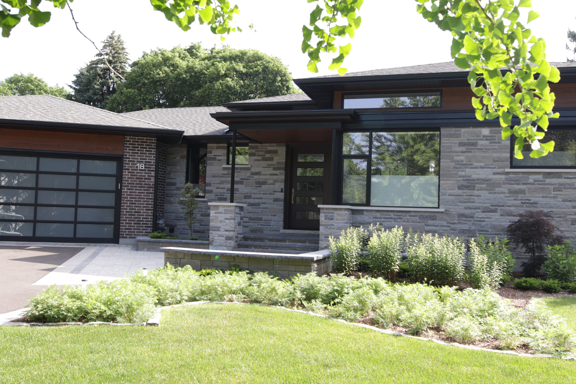 A modern house with large windows, stone façade, and a dark roof features landscaped gardens, a neat lawn, and a prominent garage door.