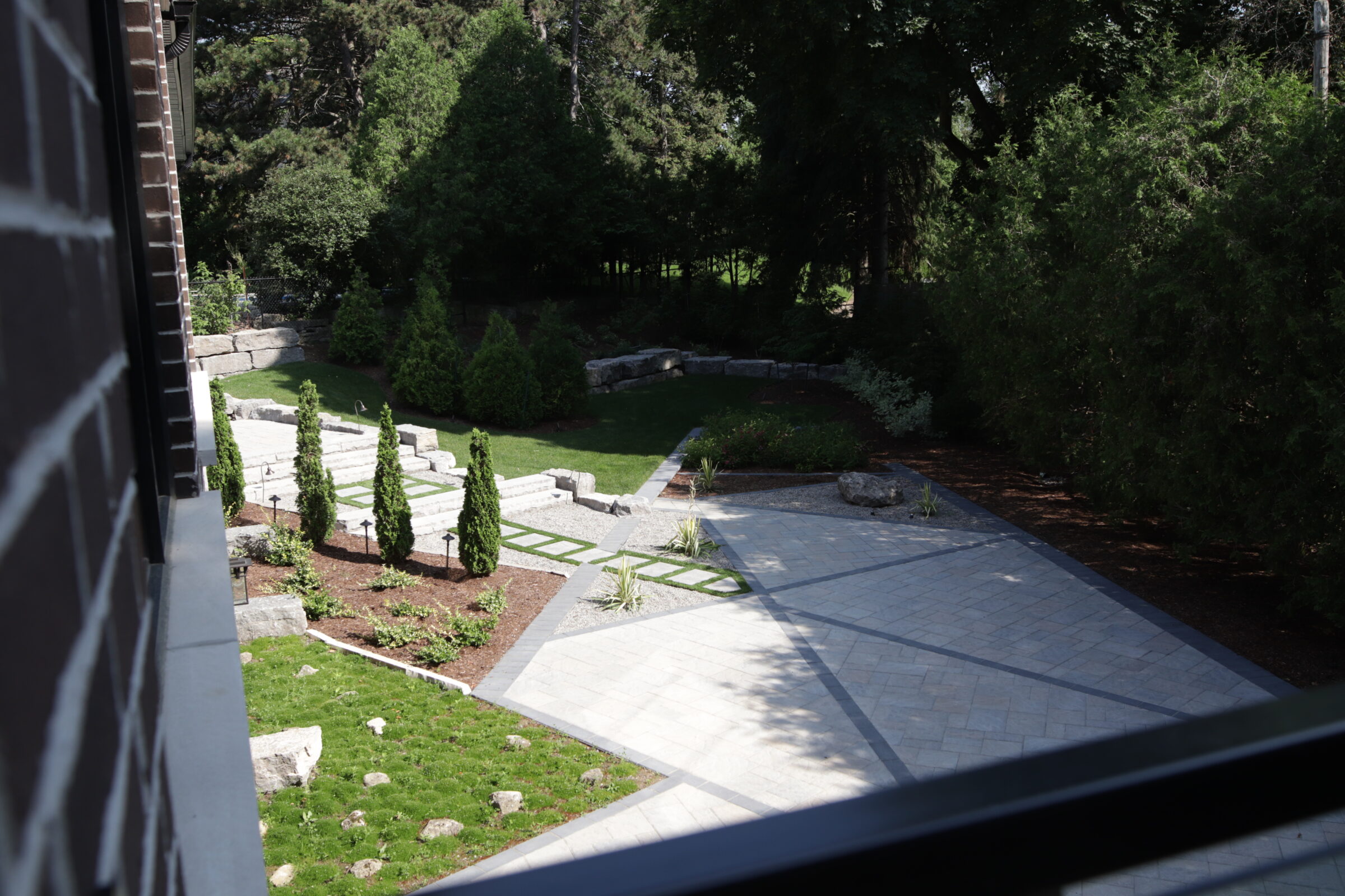 The image shows a landscaped garden with stone pathways, a lawn area, orderly shrub plantings, and trees. It's viewed from above, seemingly from a balcony.