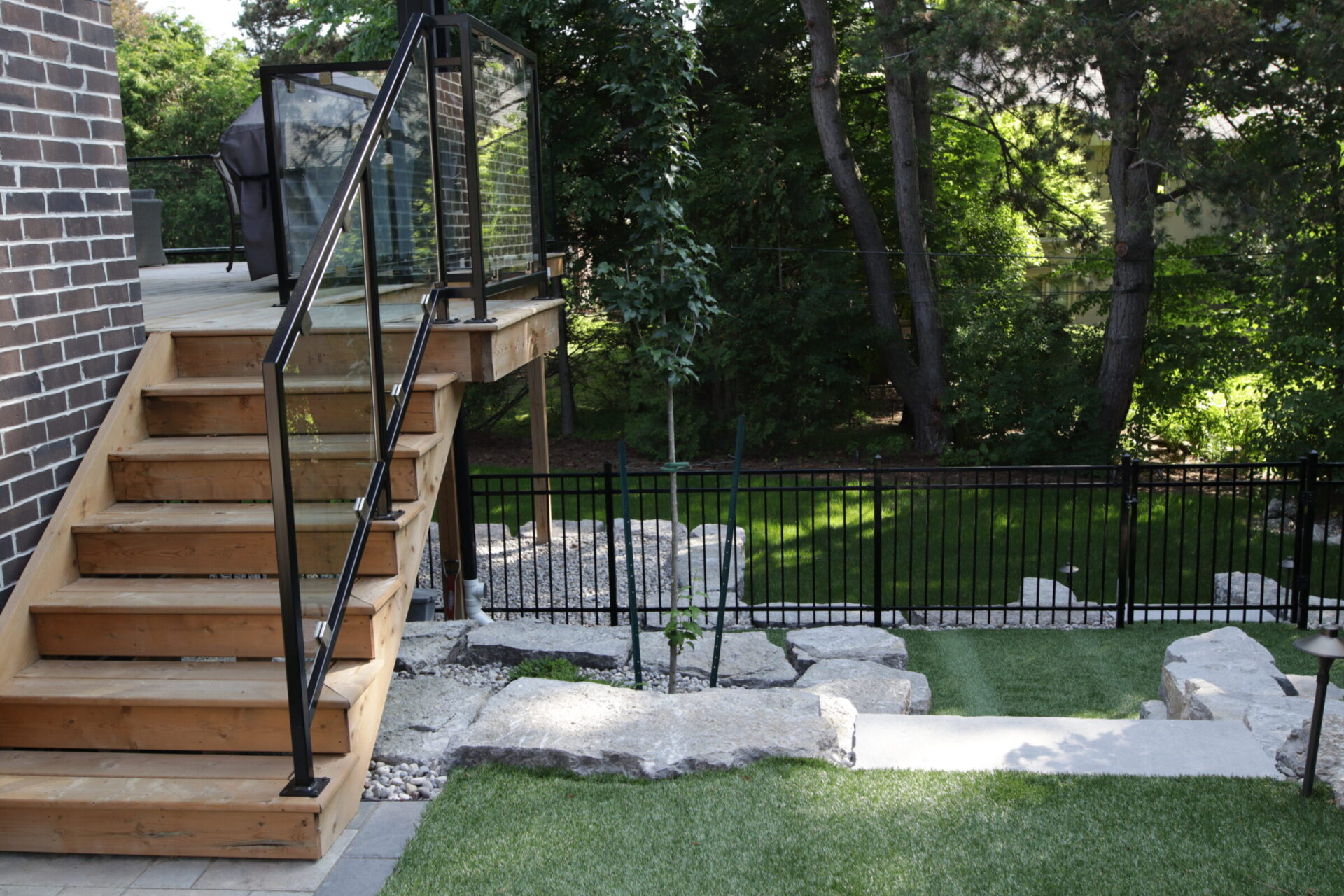 This image shows a wooden staircase with a metal railing leading to a deck. A well-kept lawn, stone landscaping, and a metal fence are visible.