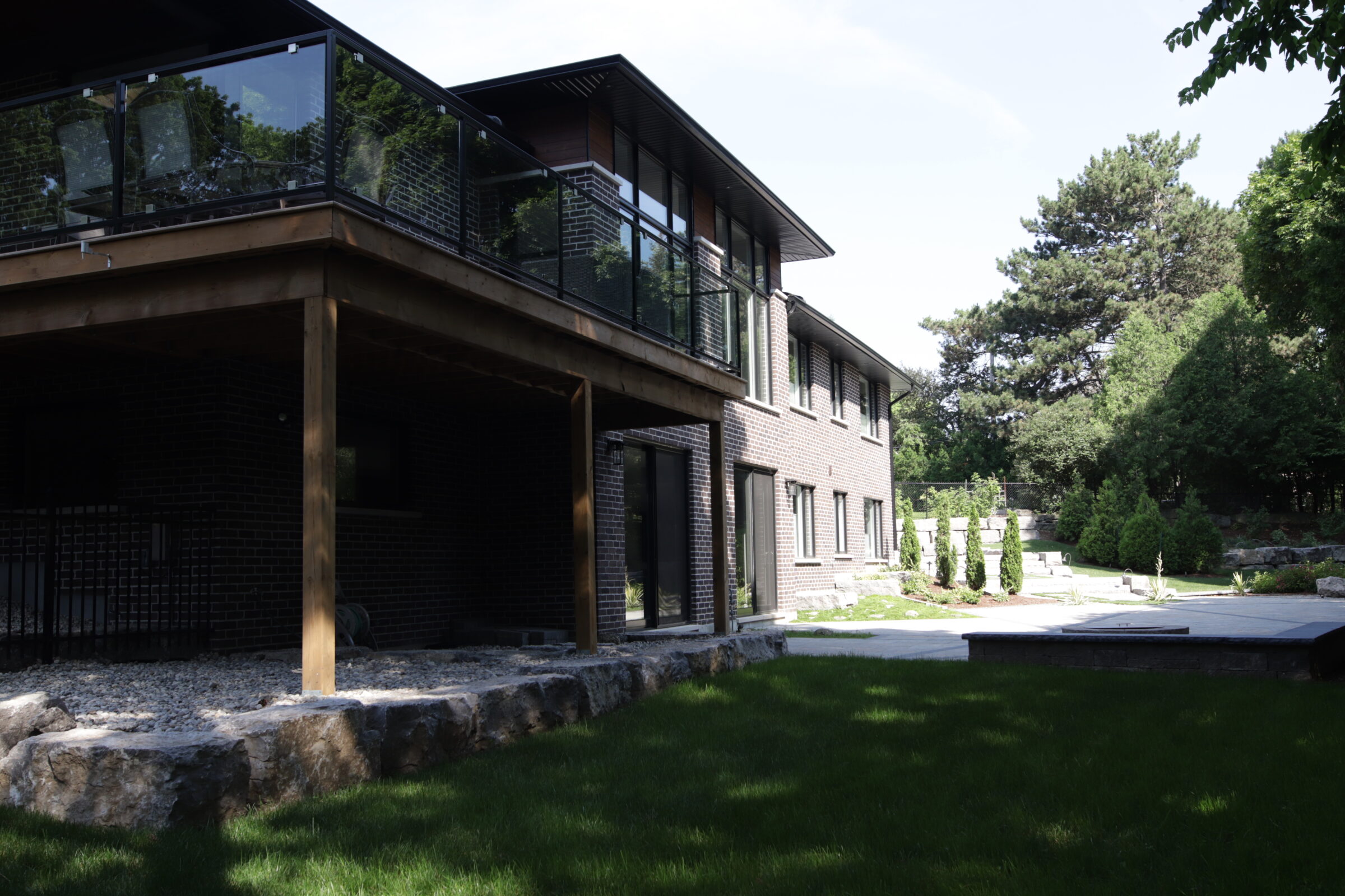 A modern two-story house with dark brick walls and a flat roof, featuring large windows, wooden pillars, and a balcony with a glass railing. Surrounded by greenery and a neatly manicured lawn.