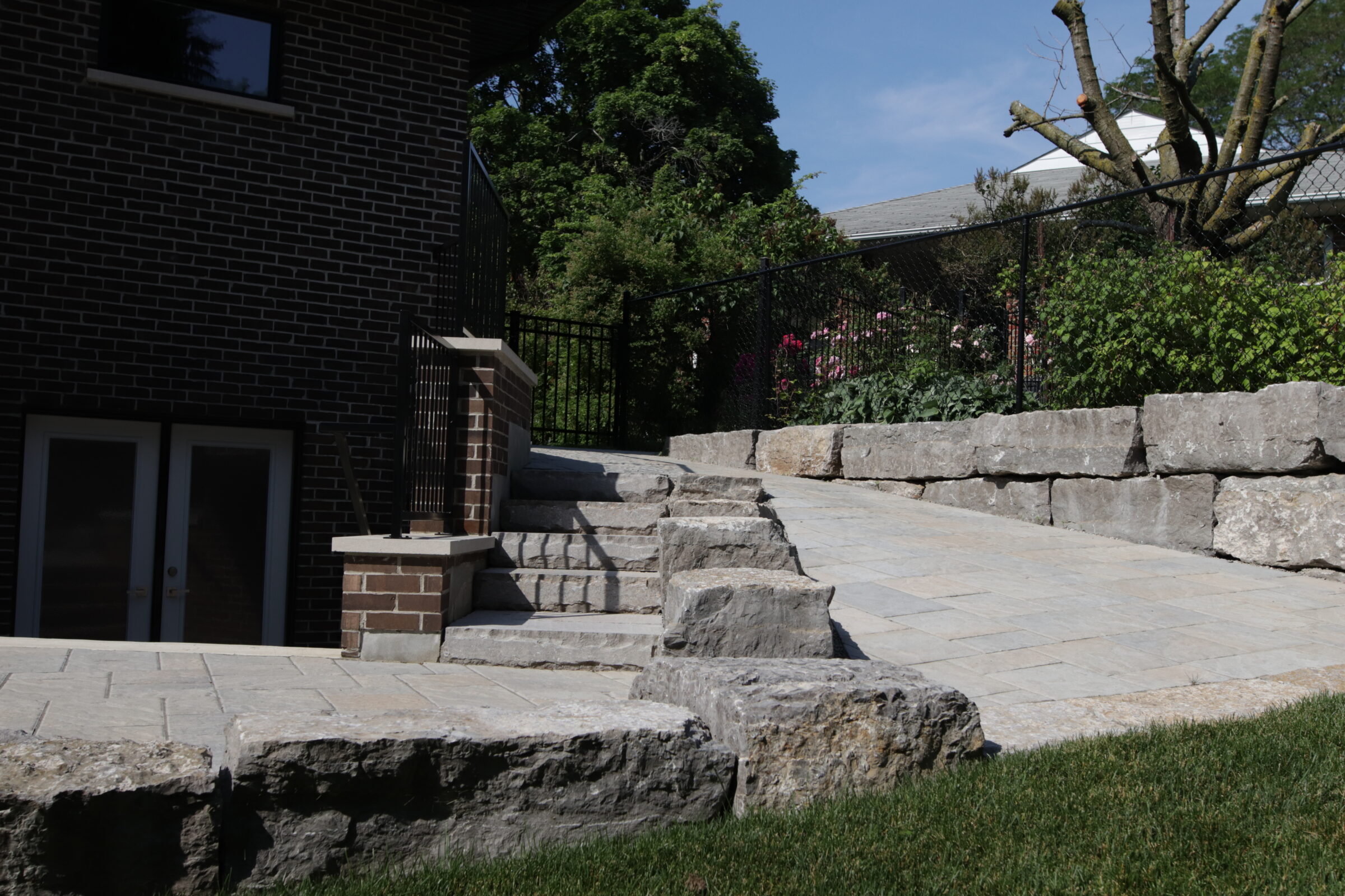 An outdoor scene featuring a stone staircase leading up to a terrace beside a brick building, with greenery and a metal fence in the background.