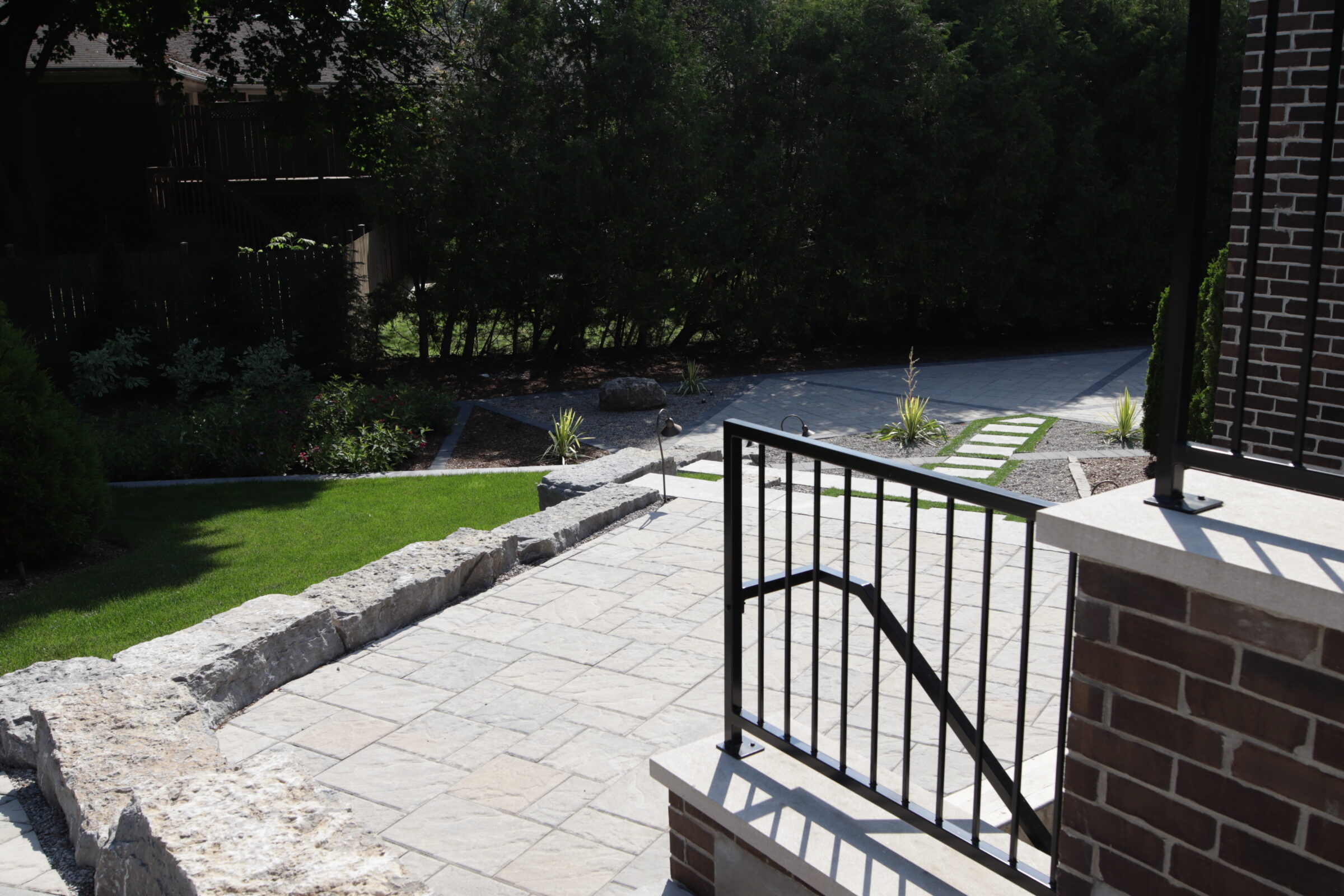 A well-manicured garden with a stone patio and pathway, lush grass, decorative plants, and a small iron gate leading to a residential property.