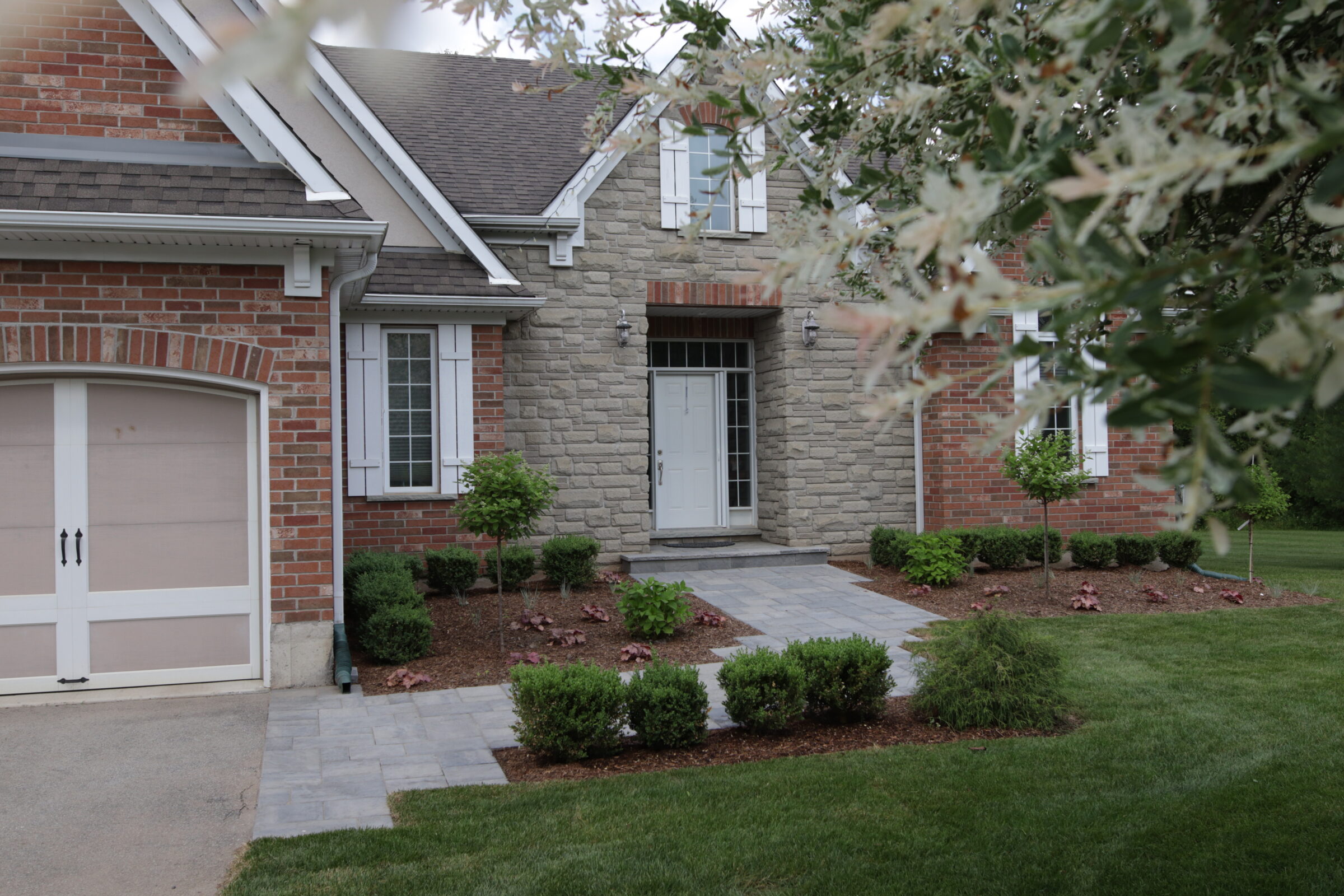 A suburban house featuring a garage, stone and brick facade, front door with side windows, walkway, landscaped garden, and a foreground of blurred leaves.