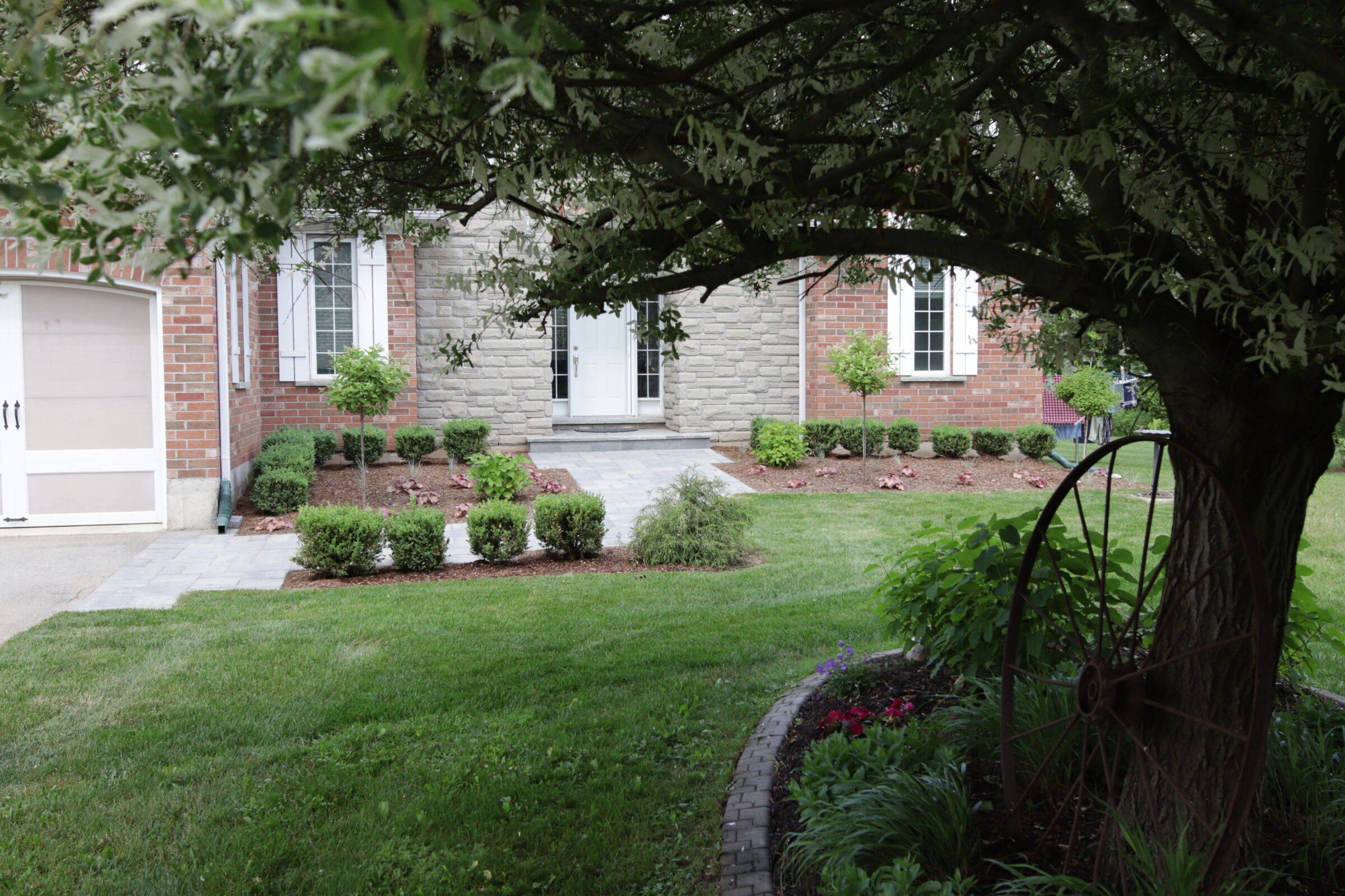 A lush green front yard of a brick house with well-manicured landscaping, pathway, stone accents, and a garage under an overcast sky.