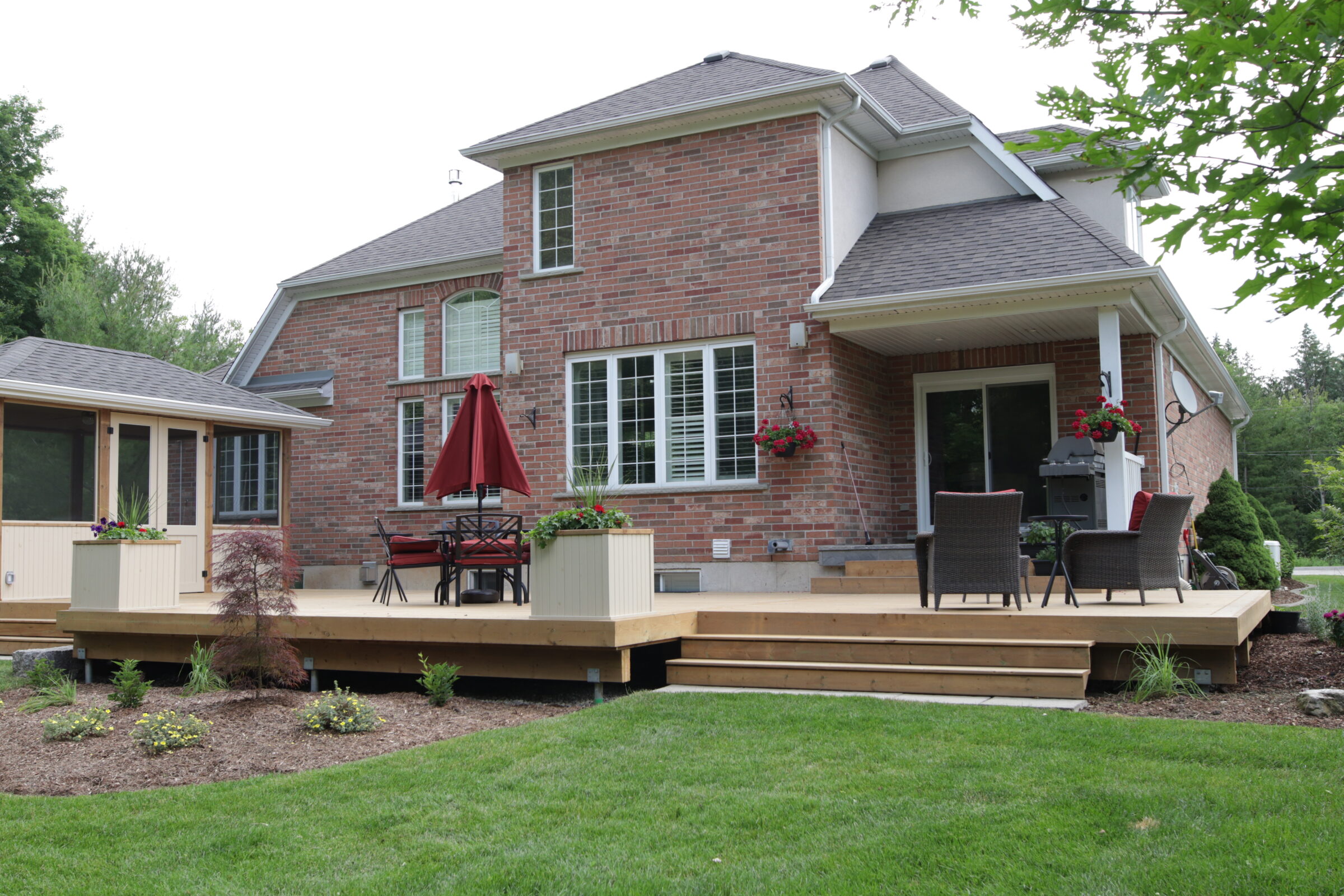 This image shows a two-story brick house with a wooden deck, outdoor furniture, potted plants, and a well-manicured lawn with young landscaping.