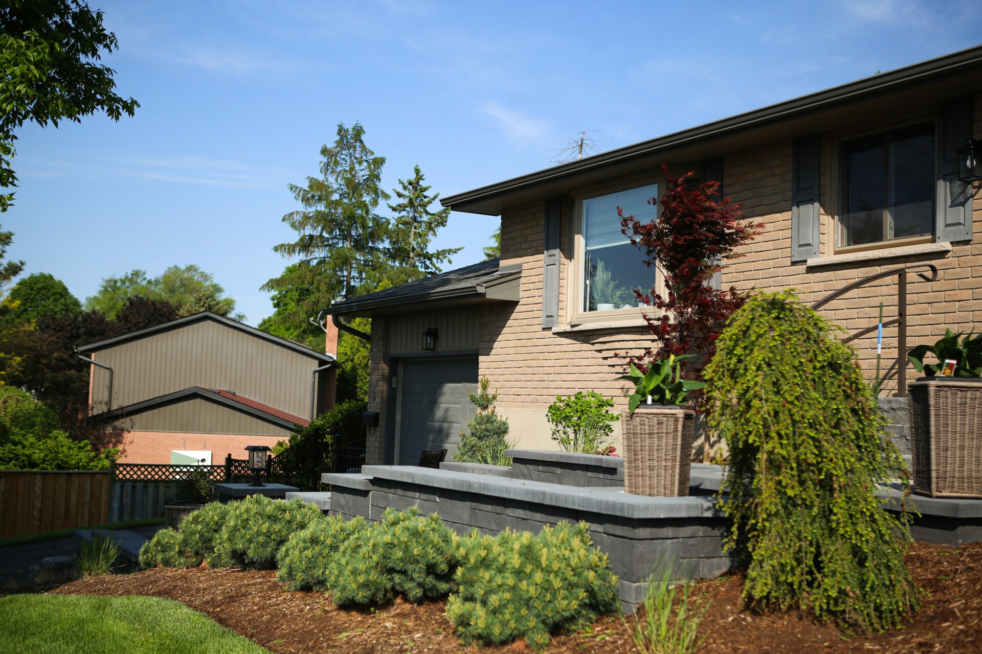 A split-level suburban house with a brick facade and a landscaped yard featuring shrubs, trees, and a neatly trimmed lawn under a blue sky.