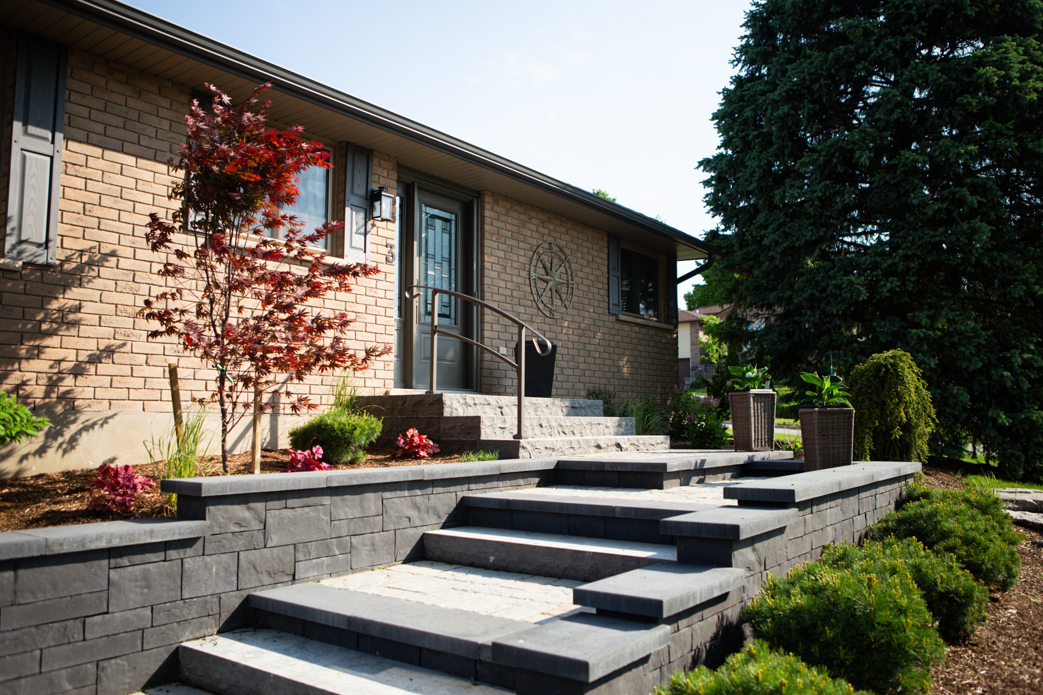 A neat residential brick home with a landscaped front yard featuring stone stairs, potted plants, shrubs, and a small red tree under a sunny sky.
