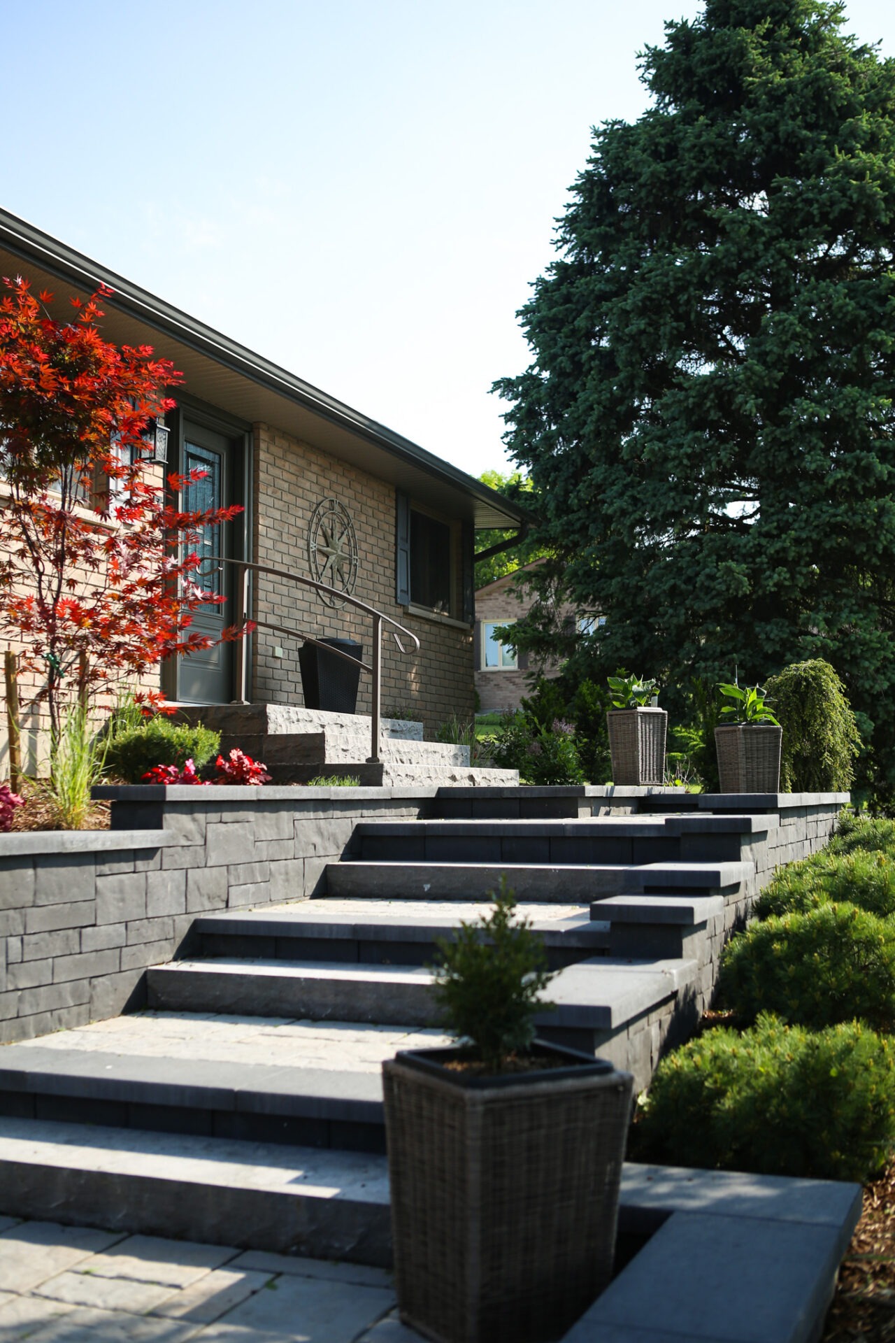 A brick house with stylish dark stone steps leading to the entrance, flanked by plants, under a clear blue sky. A large conifer stands nearby.