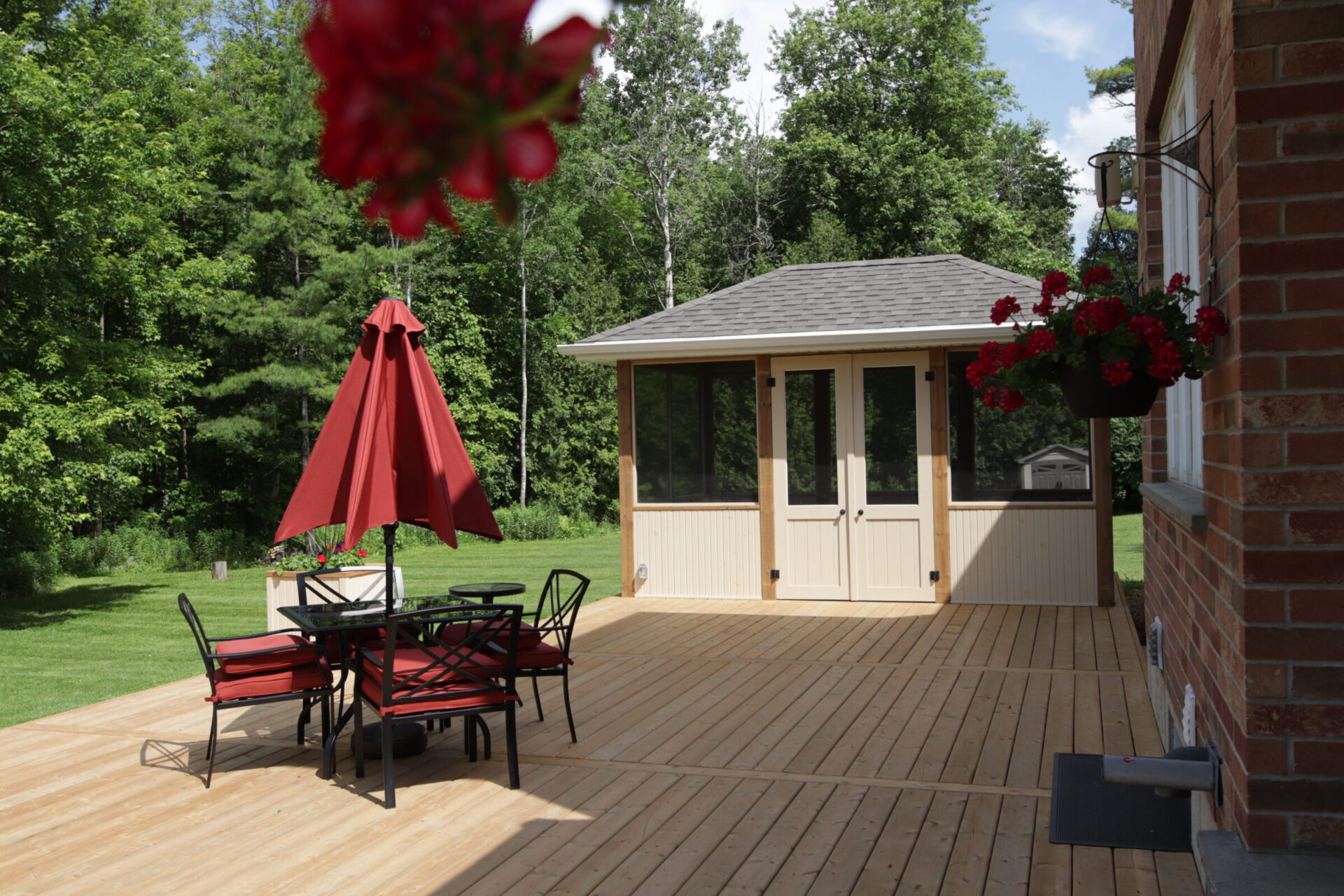 A spacious wooden deck with a red patio umbrella and outdoor dining furniture, adjacent to a small gazebo, with lush greenery in the background.