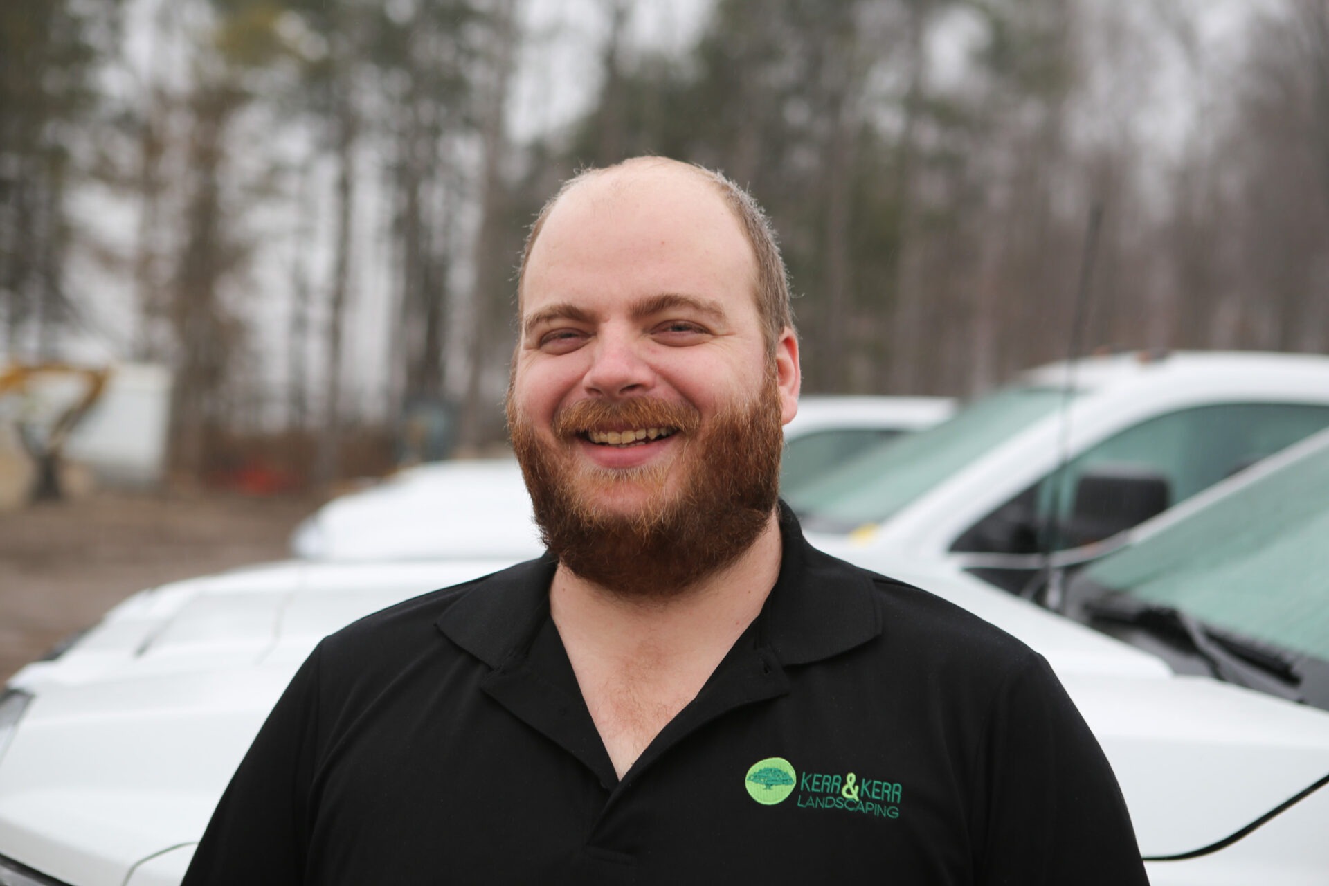 A person with a beard is smiling in front of vehicles, wearing a black shirt with a landscaping company logo, in a blurred background setting.
