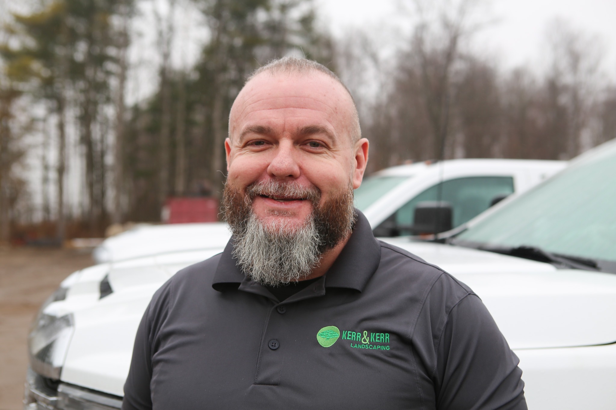 A person with a short haircut and a full beard is smiling at the camera, wearing a black shirt with a landscaping company logo, outdoors.