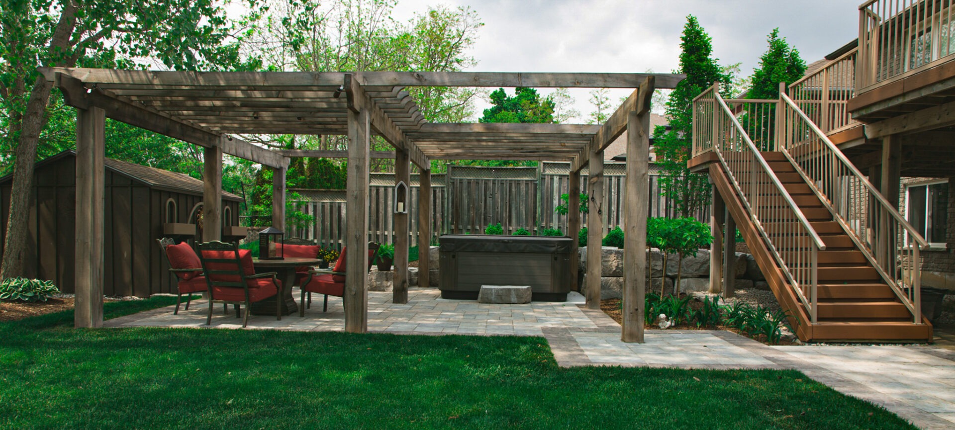 An inviting backyard with a wooden pergola, outdoor furniture with red cushions, stone patio, neatly manicured lawn, and a set of stairs leading up a deck.