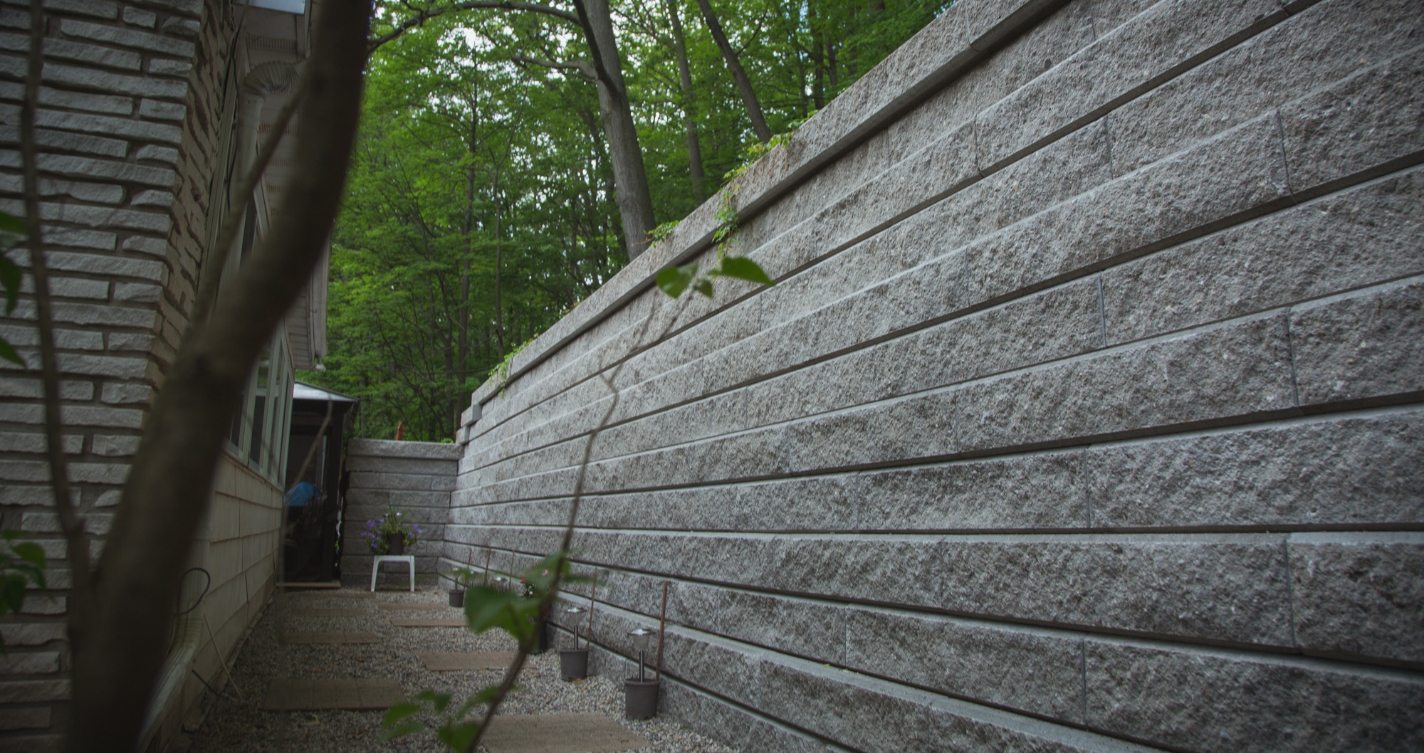 The image shows an outdoor area with a textured stone wall on the right and a building on the left, amidst a backdrop of green trees.