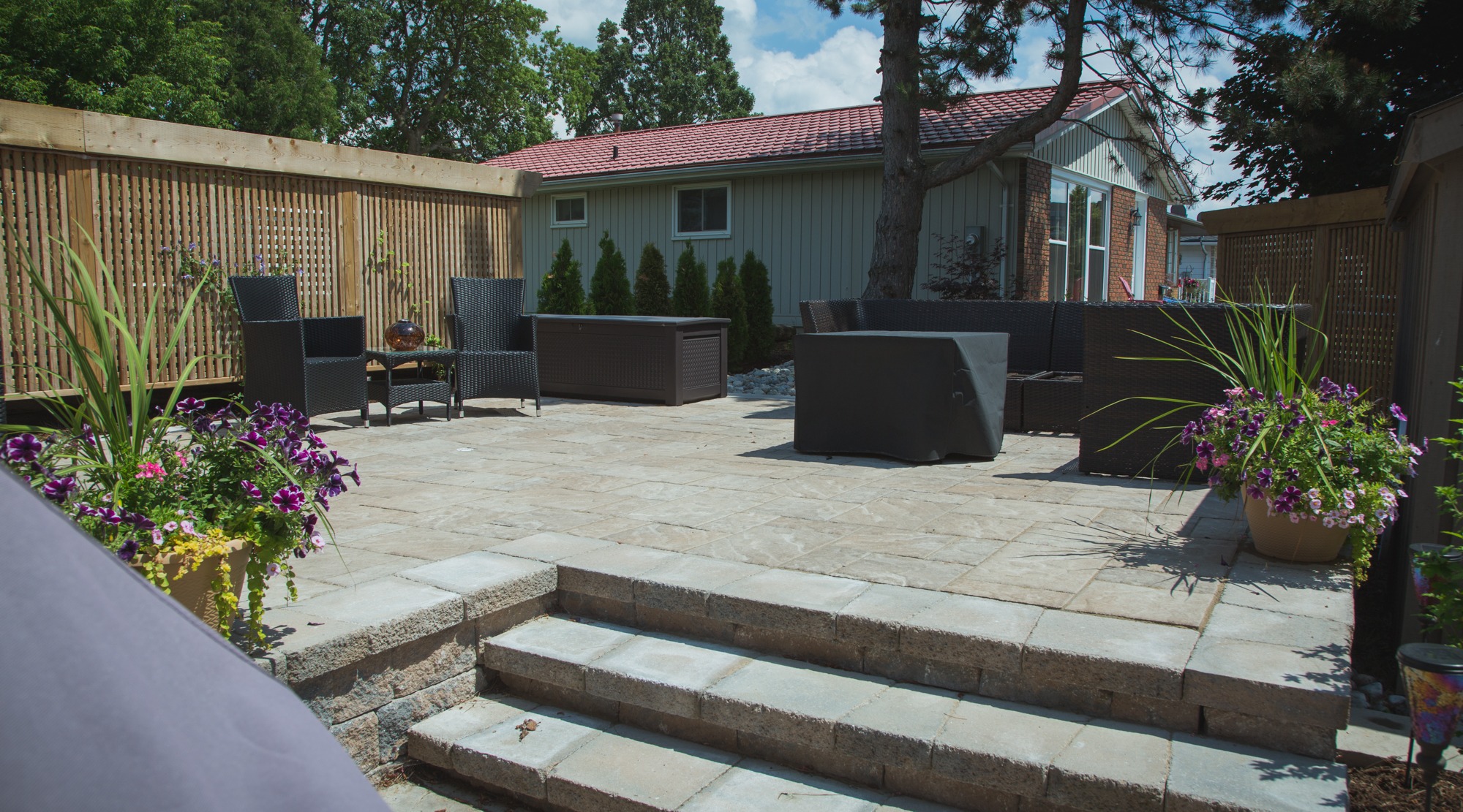 A sunny backyard patio with stone tiles, surrounded by a wooden fence, furnished with a black wicker seating set, potted plants, and a grill covered.