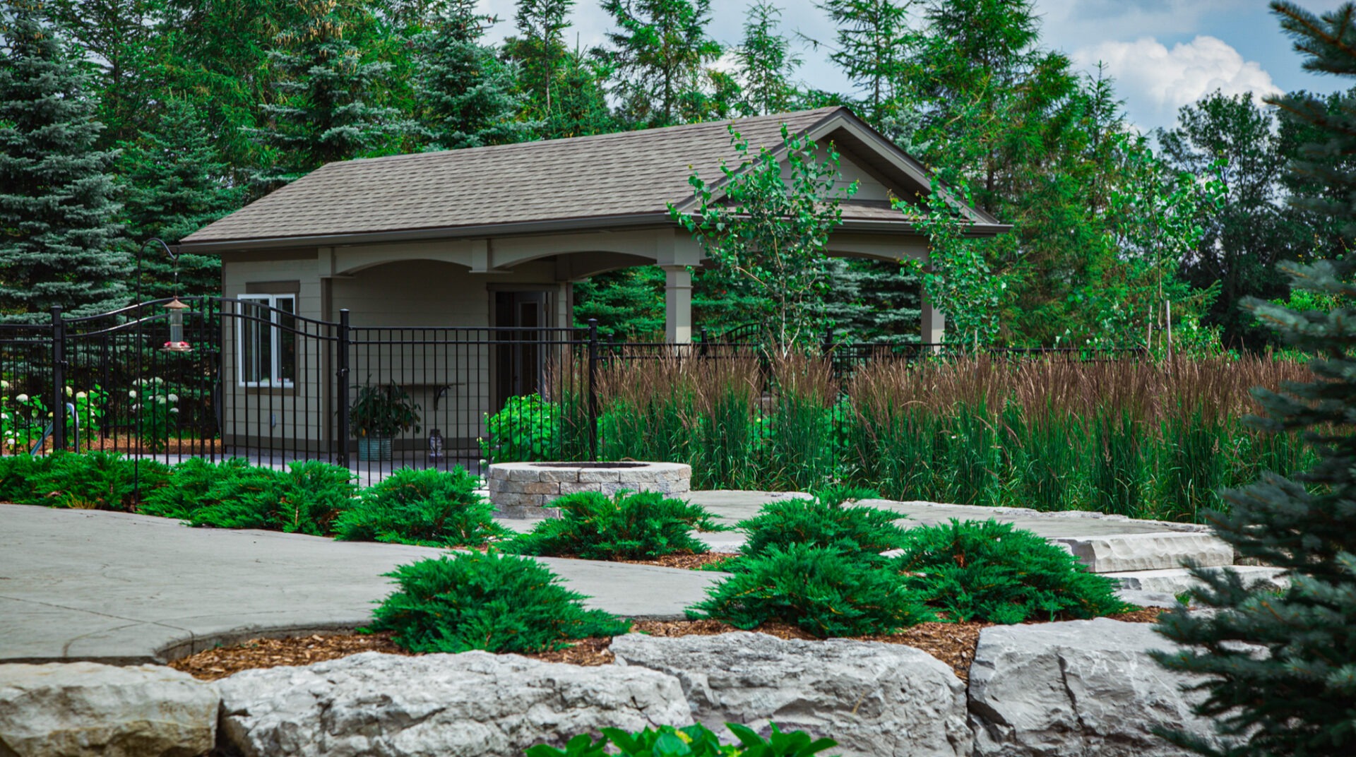 A small beige building with a hip roof is nestled amongst lush greenery and trees, bordered by a black metal fence and stone pathway.