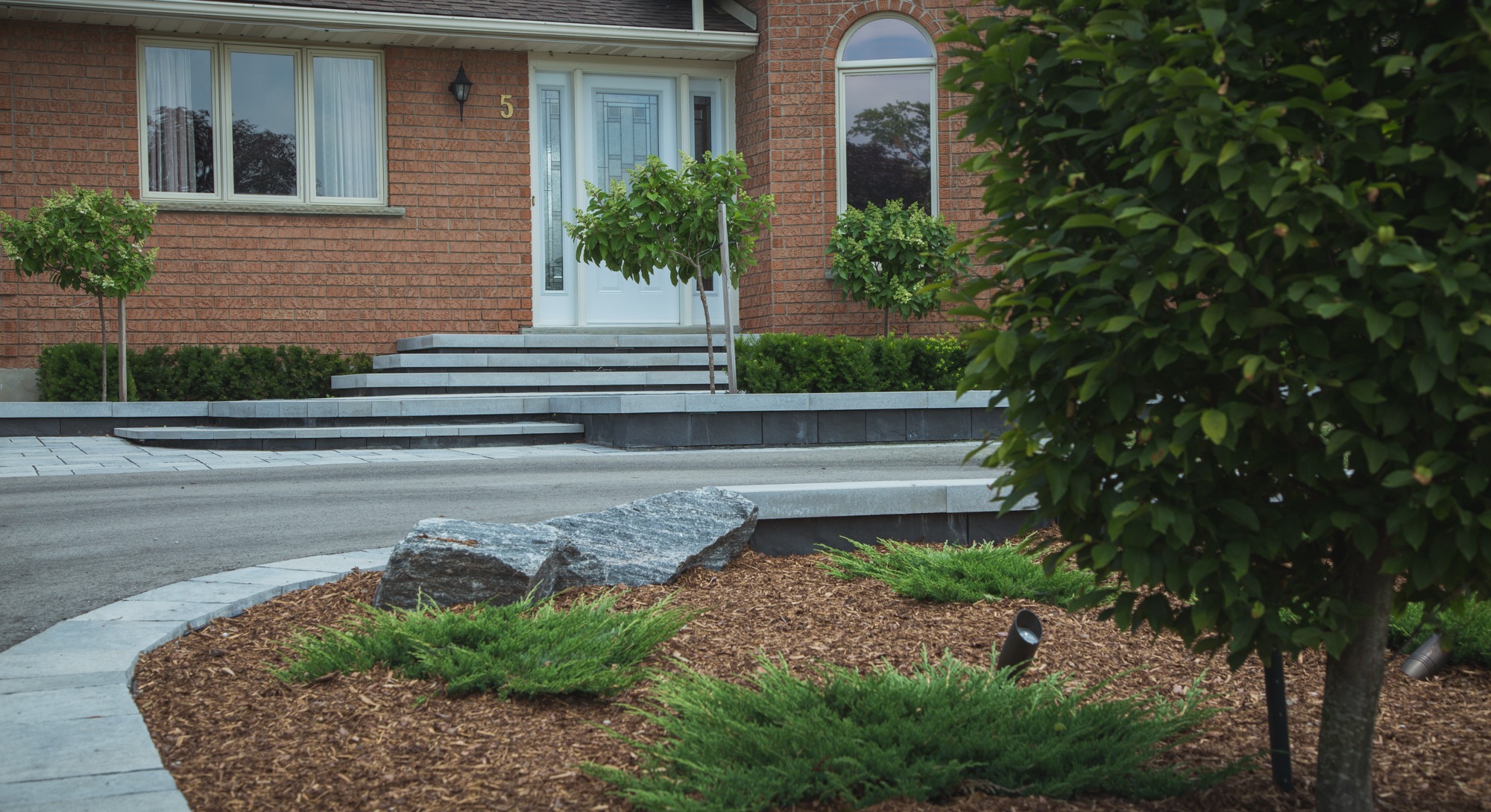 A well-maintained front yard with tidy landscaping, mulch, shrubs, a young tree, and a brick house with a number 5 on its facade.