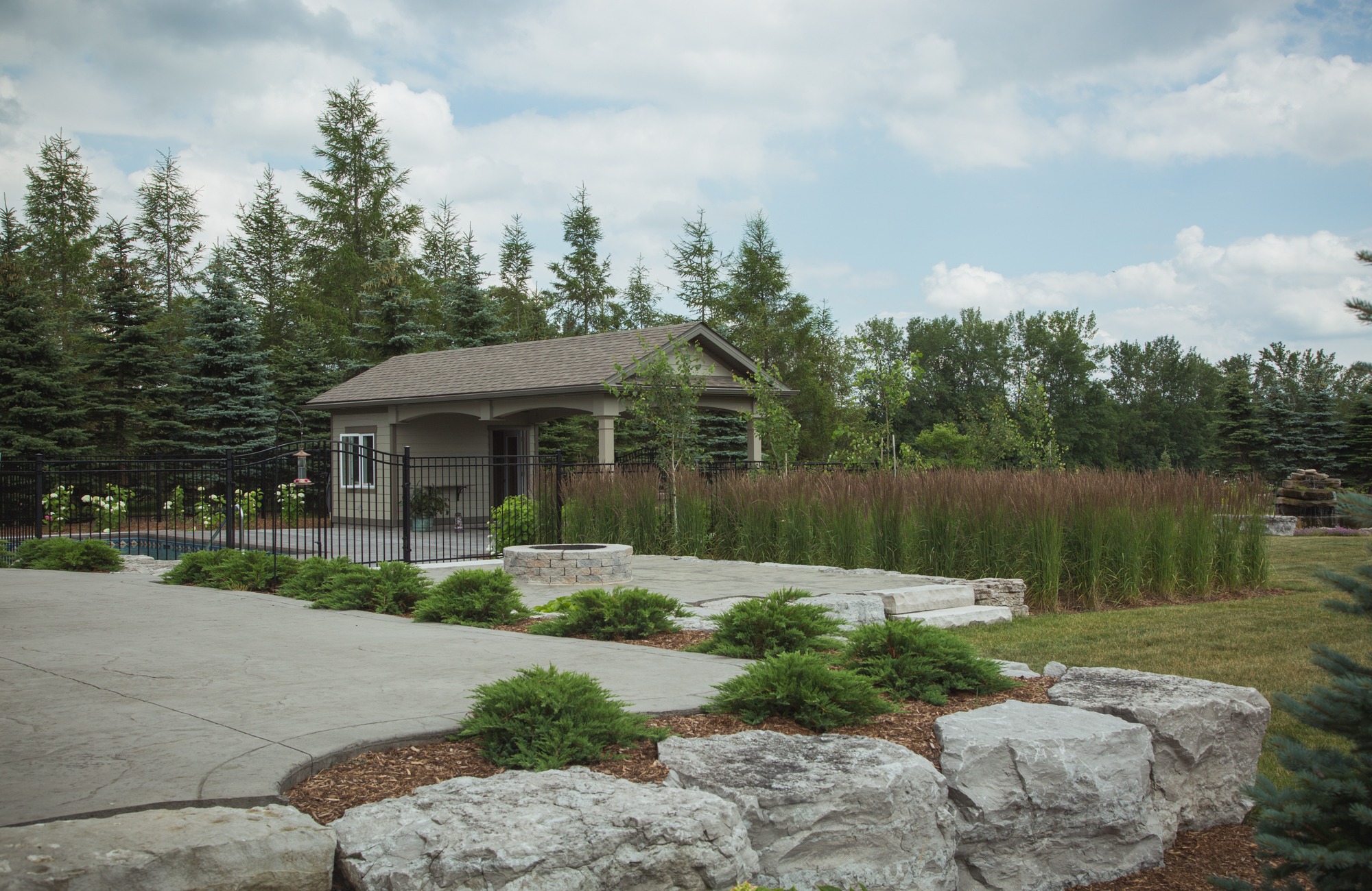 This image shows a small gatehouse at the entrance to a park or property flanked by a black metal gate, landscaped with stones and greenery.
