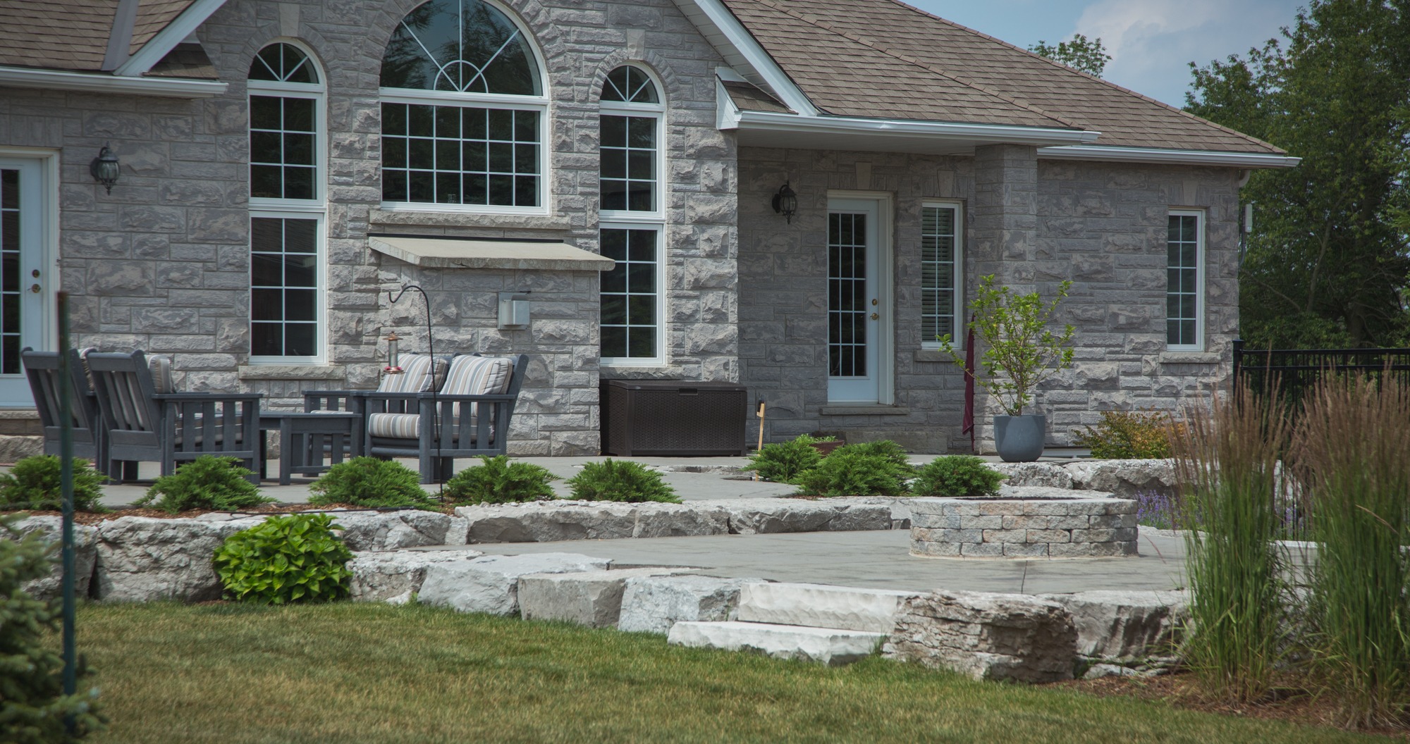 This image shows a stone house with arched windows, a patio with outdoor furniture, and a landscaped garden with stone steps leading up to it.