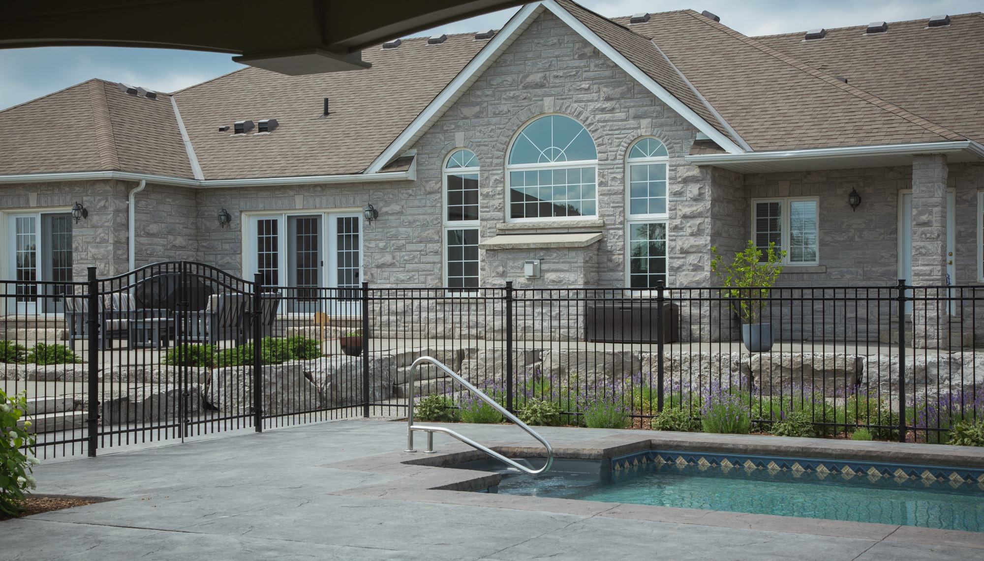An elegant stone house with large windows, a shingle roof, and a fenced outdoor area featuring a swimming pool with a metal ladder.