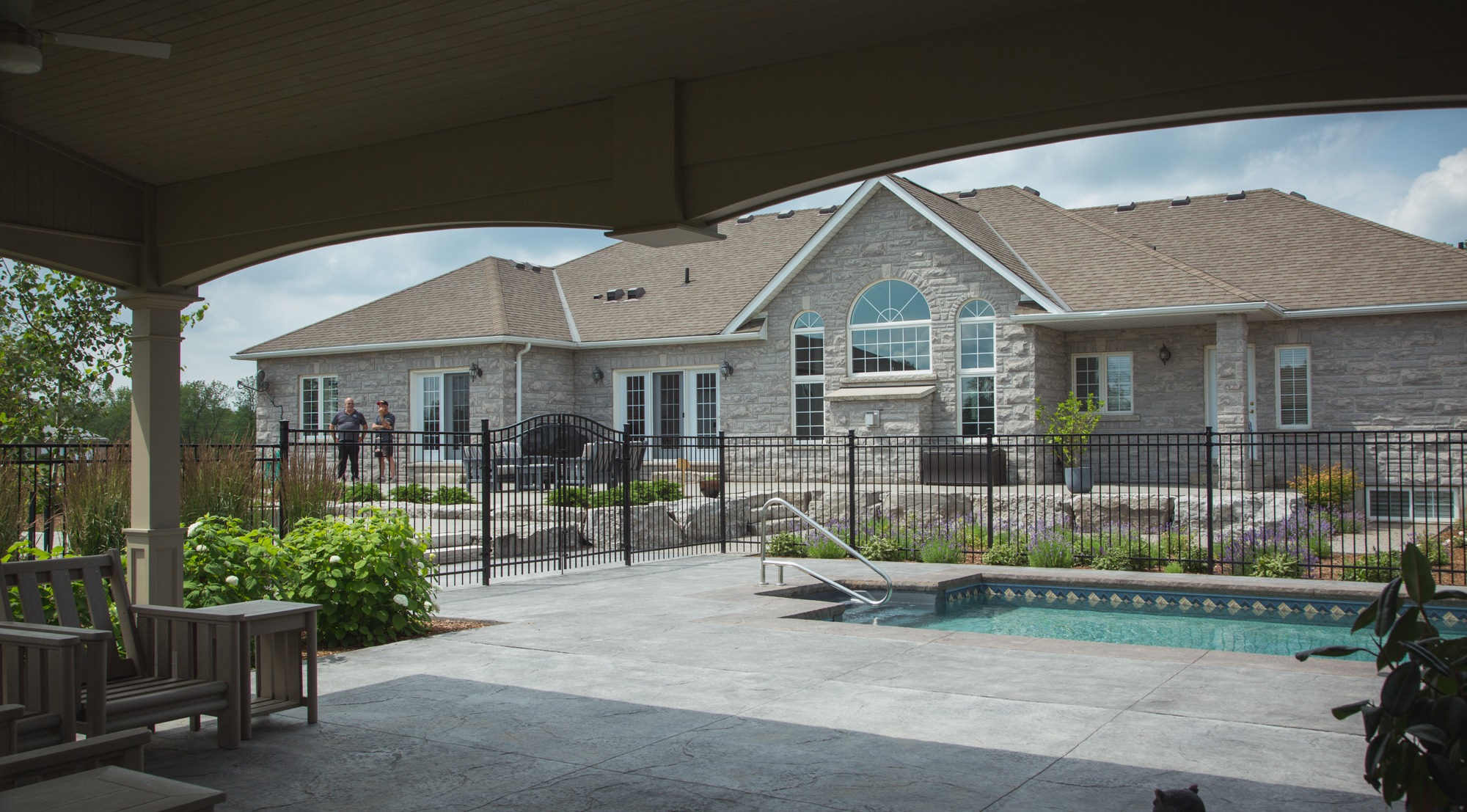 The image shows a stone house with large windows, a covered patio with seating, and a fenced pool area. Two people stand by the railing, chatting.