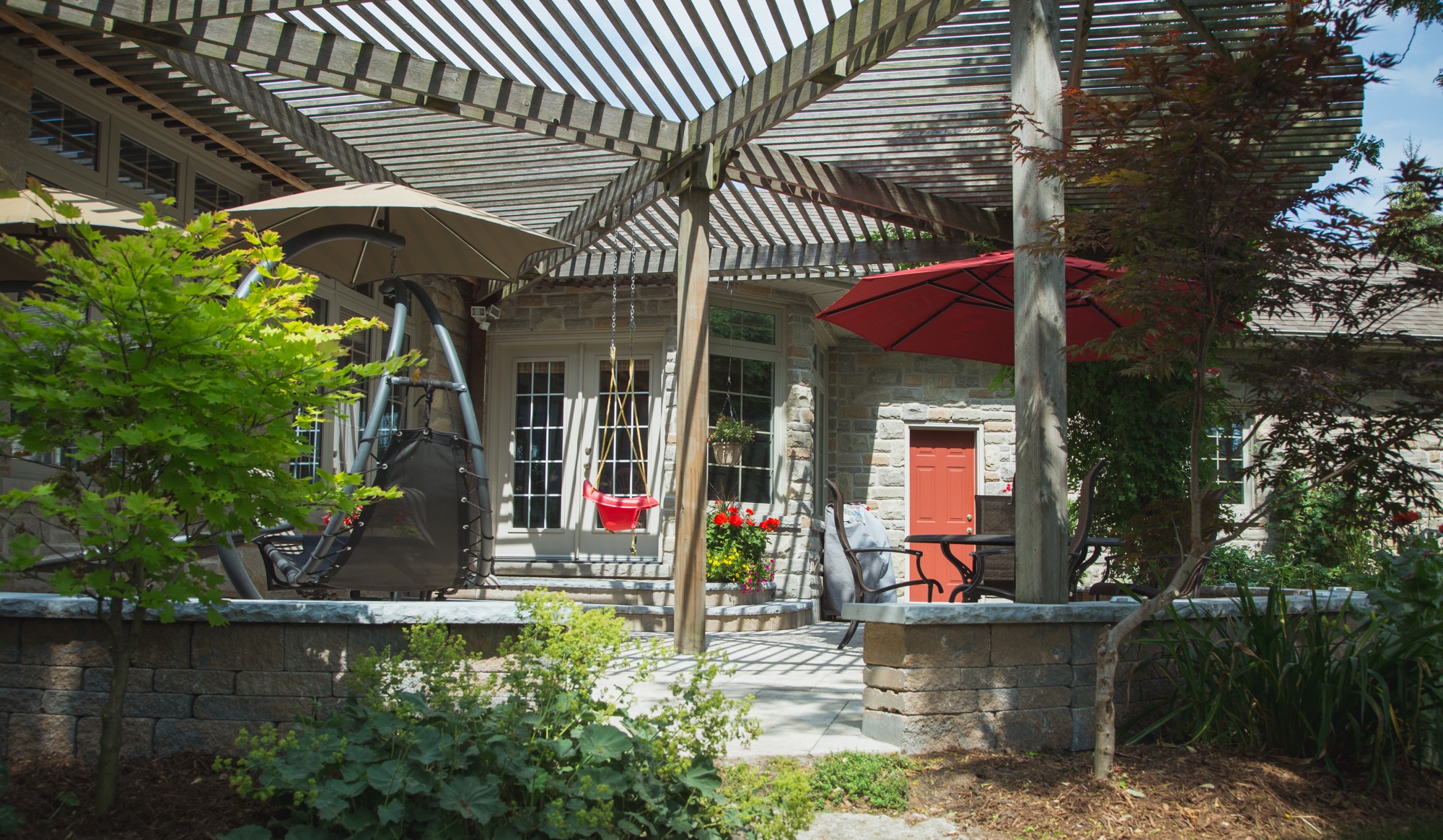 A cozy patio area with a wooden pergola, stone house, red accents on doors and umbrella, hanging chair, and lush greenery on a sunny day.