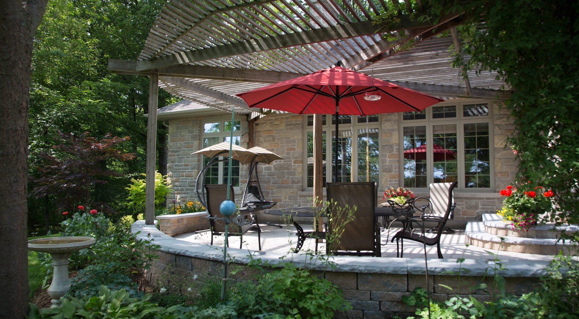 A serene patio with a red umbrella, chairs, and a swing, surrounded by lush greenery and colorful flowers, adjacent to a stone house.