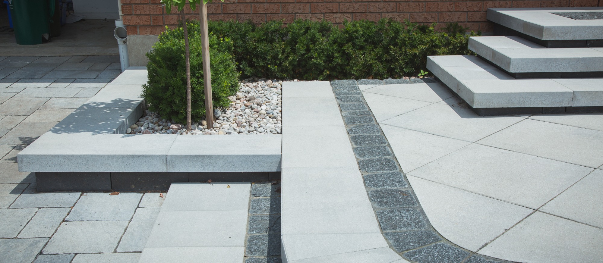 This image features a modern pathway and steps with contrasting materials, surrounded by landscaping with small shrubs, gravel, and a young tree.