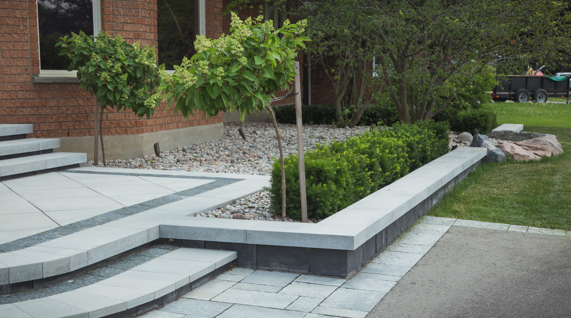 A landscaped garden with geometric stepping stones, a raised bed, young trees, shrubs, and a mix of grass and gravel by a brick building.