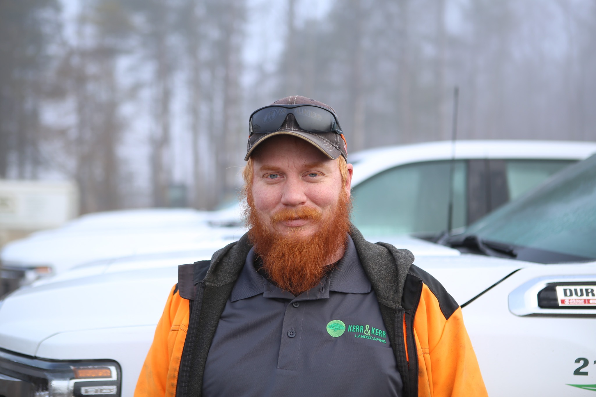 A person with a red beard and baseball cap is standing in front of a white vehicle with a blurred forest background in misty weather.