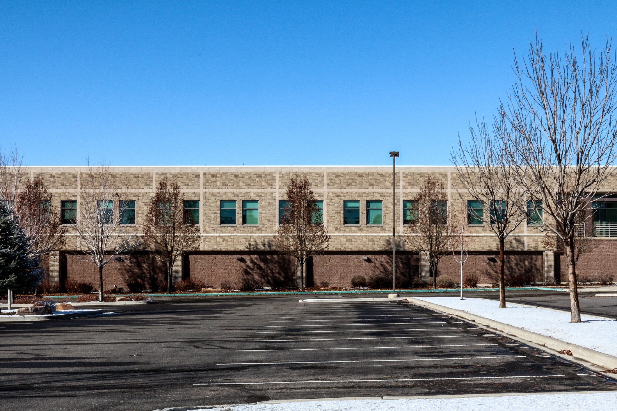 This image shows an empty parking lot in front of a modern two-story stone building with a clear blue sky and several bare trees.